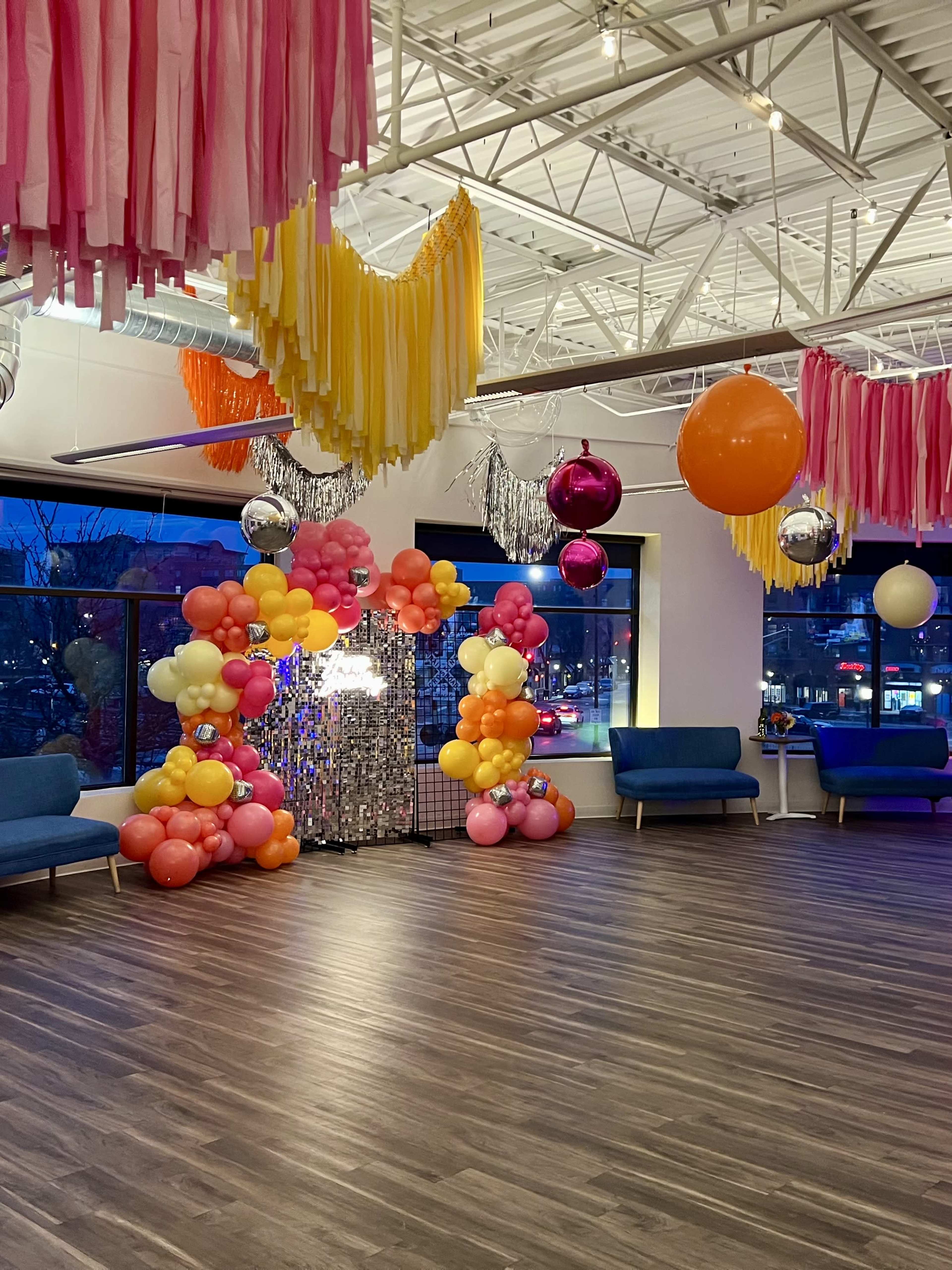 The image shows a decorated indoor event space featuring colorful balloons, streamers, and seating arrangements under bright lighting.