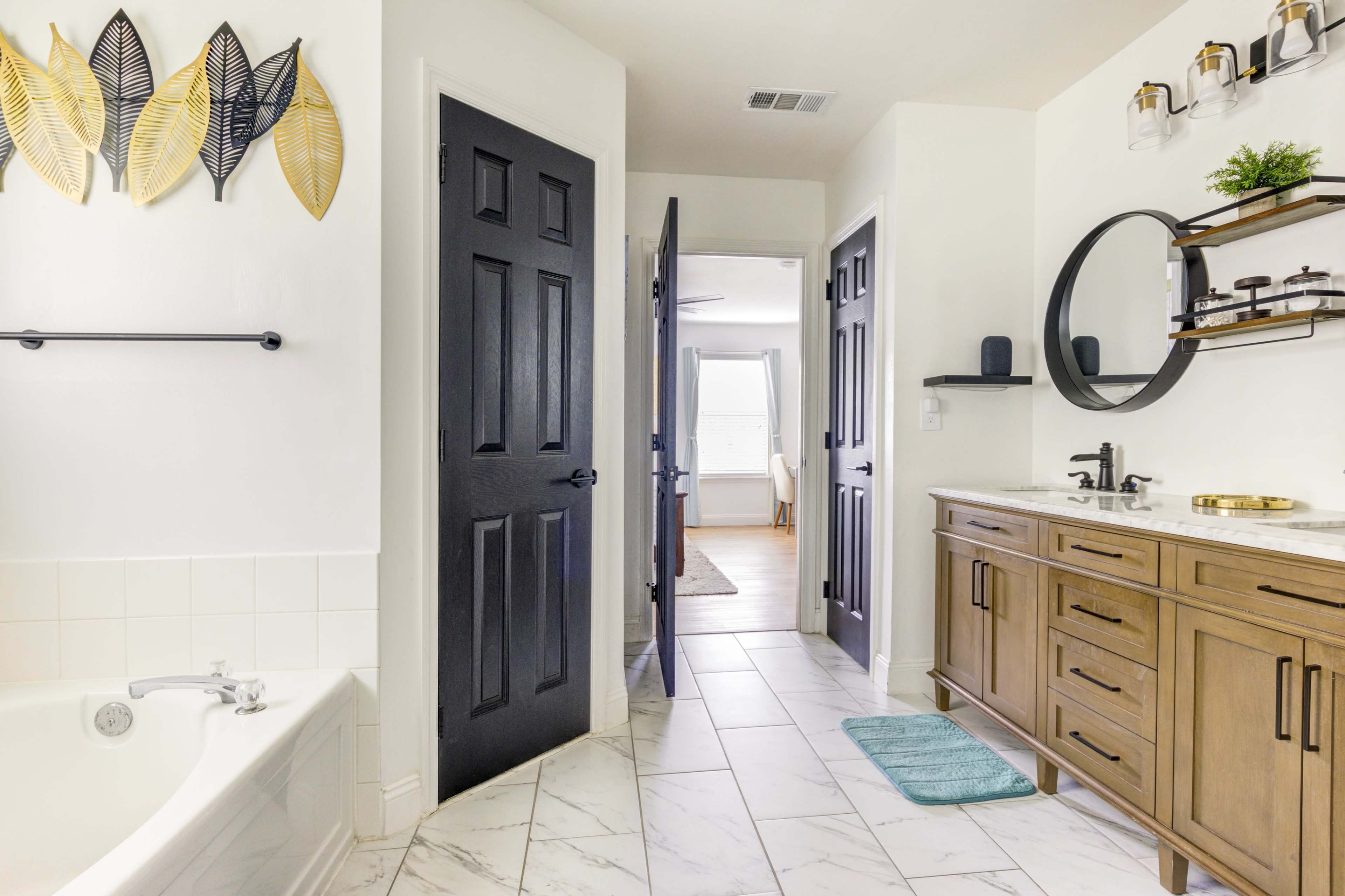 A modern bathroom with a bathtub, wooden vanity, dark double doors, and decorative leaf wall art.