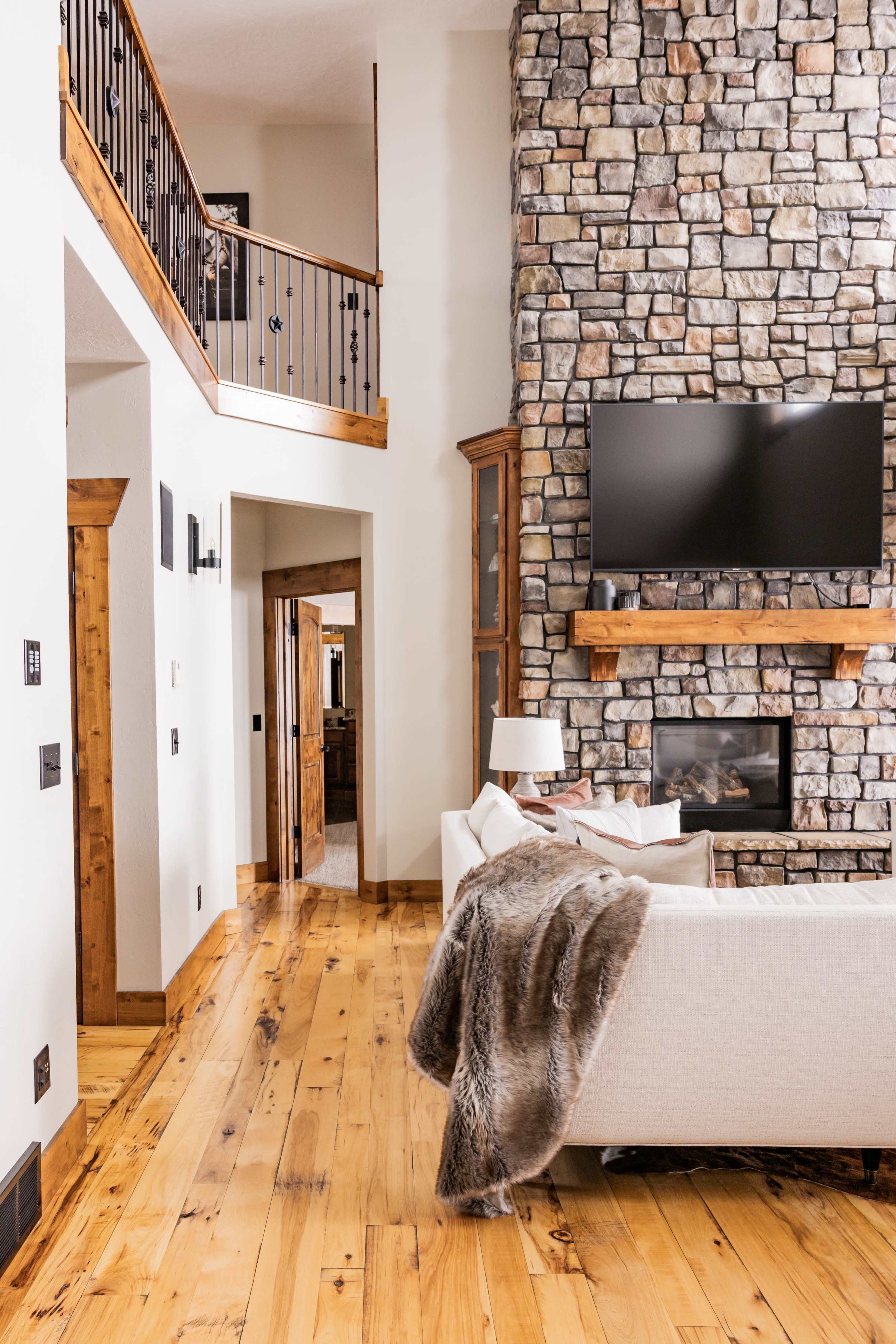 A spacious living room with a stone fireplace, hardwood floors, and a large television mounted on the wall.