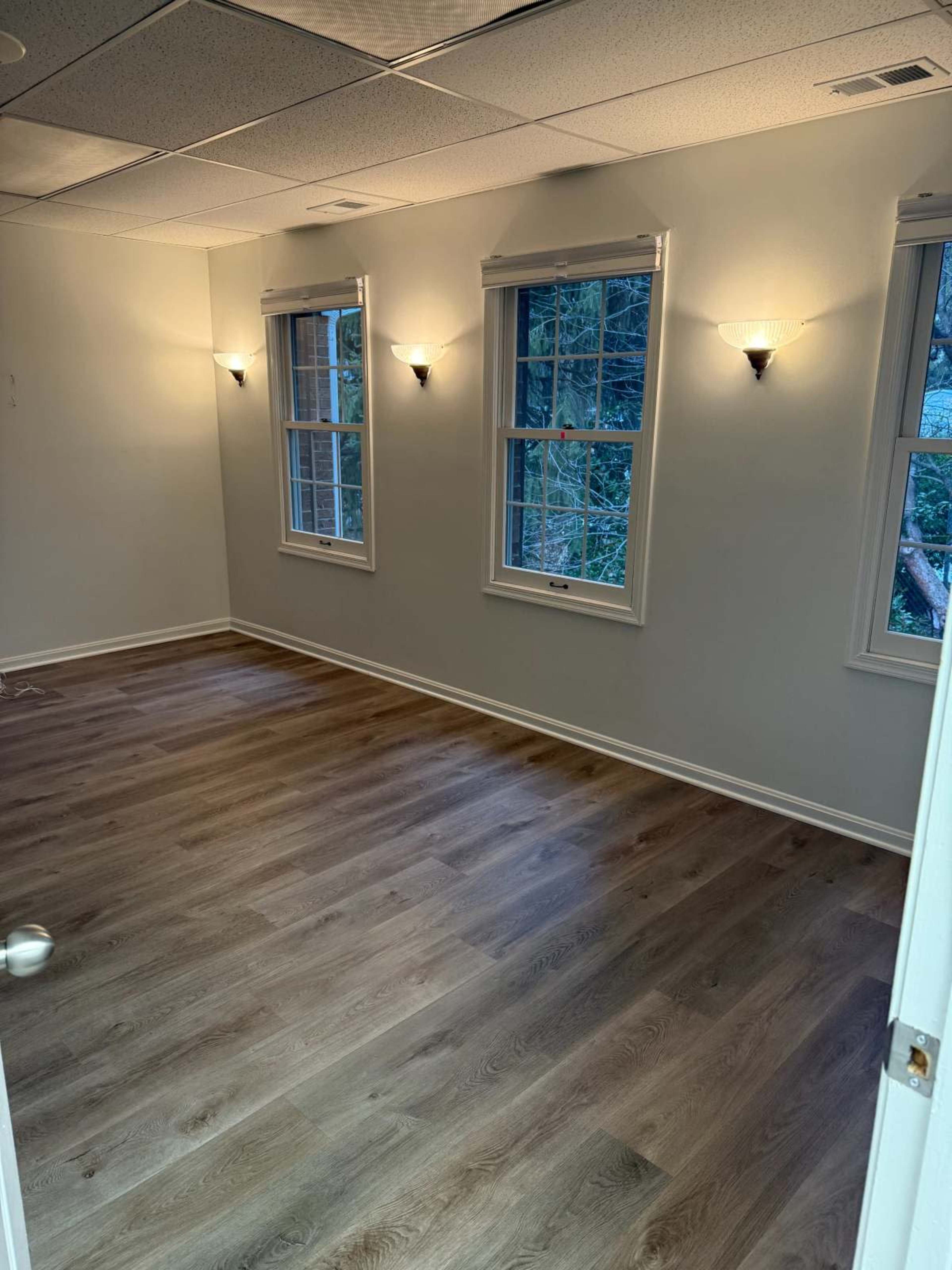 The image shows a well-lit room with wooden flooring, featuring three windows and light fixtures on the walls.