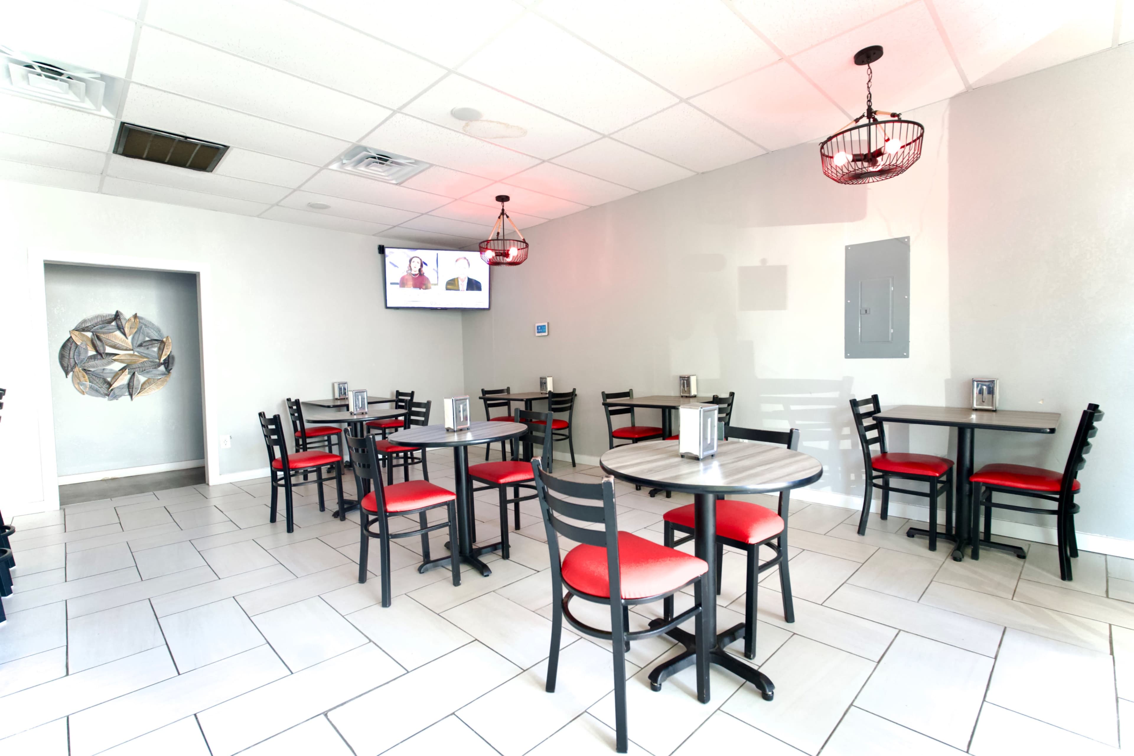 The image shows a brightly lit dining area with several tables and chairs, a television mounted on the wall, and decorative elements.