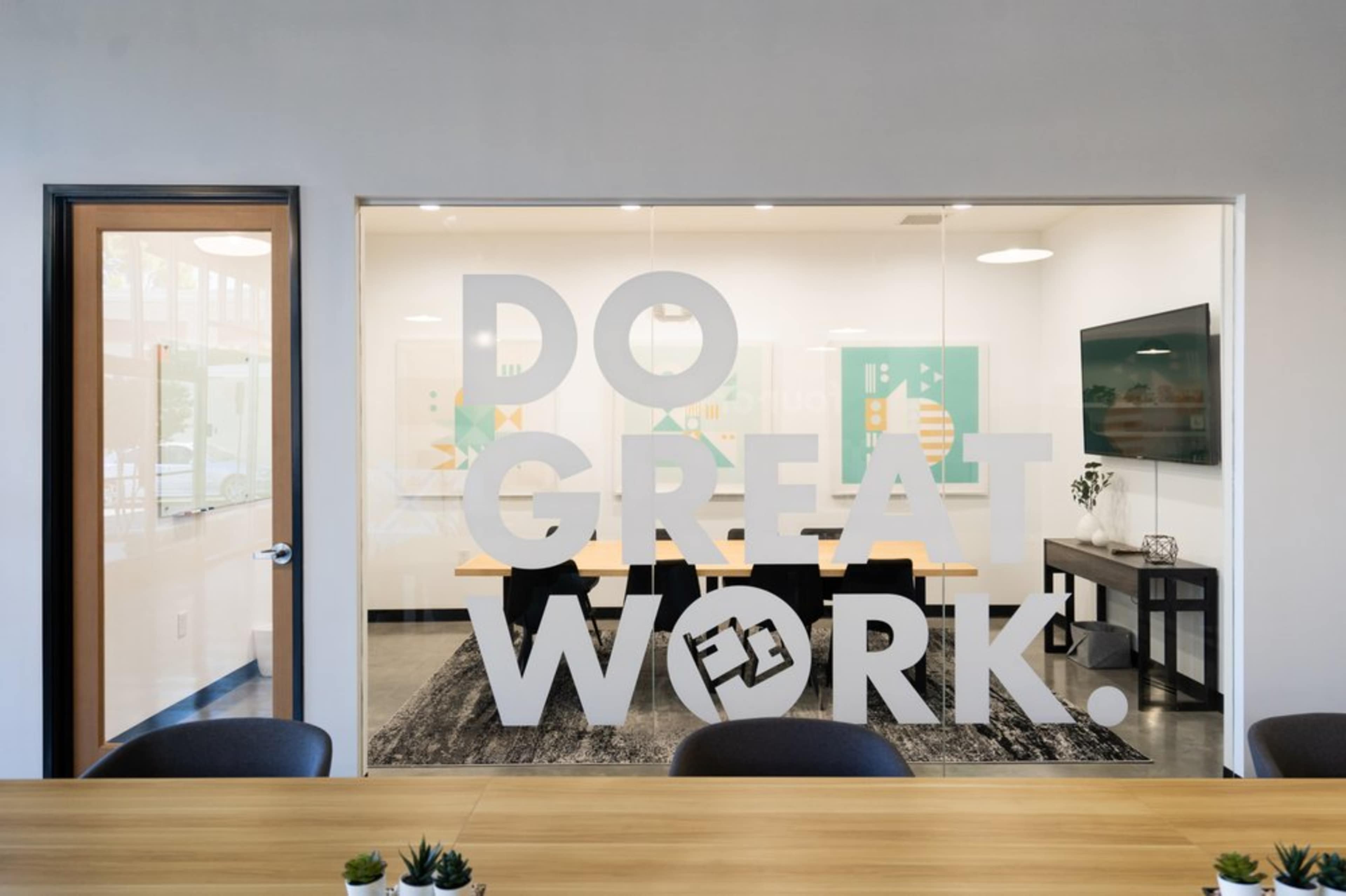 A glass-walled conference room features a modern table and chairs, with the phrase "DO GREAT WORK!" prominently displayed on the window.