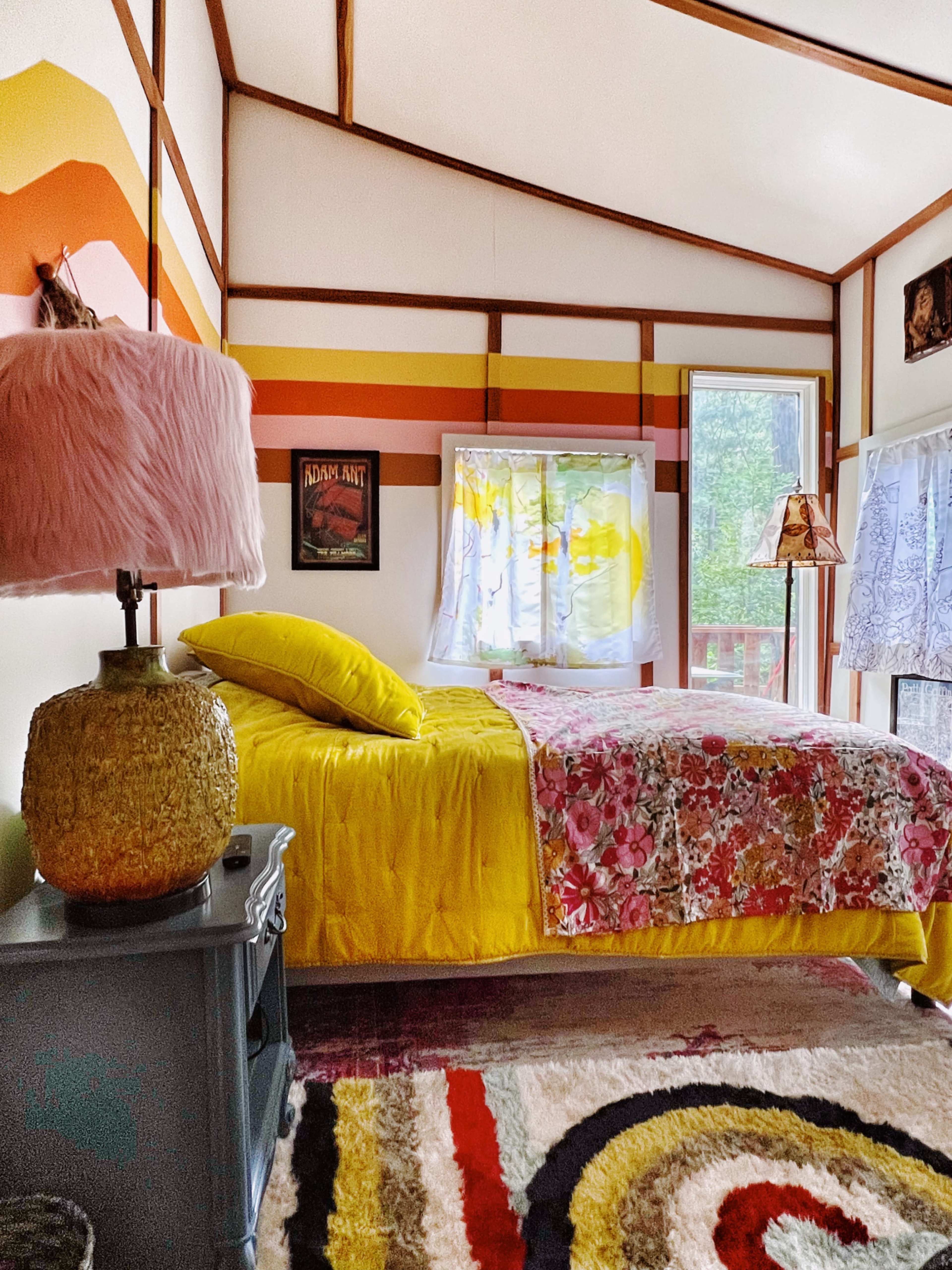 The image shows a brightly decorated bedroom featuring a yellow bedspread, patterned pillows, a colorful rug, and a mix of vintage and modern decor elements.