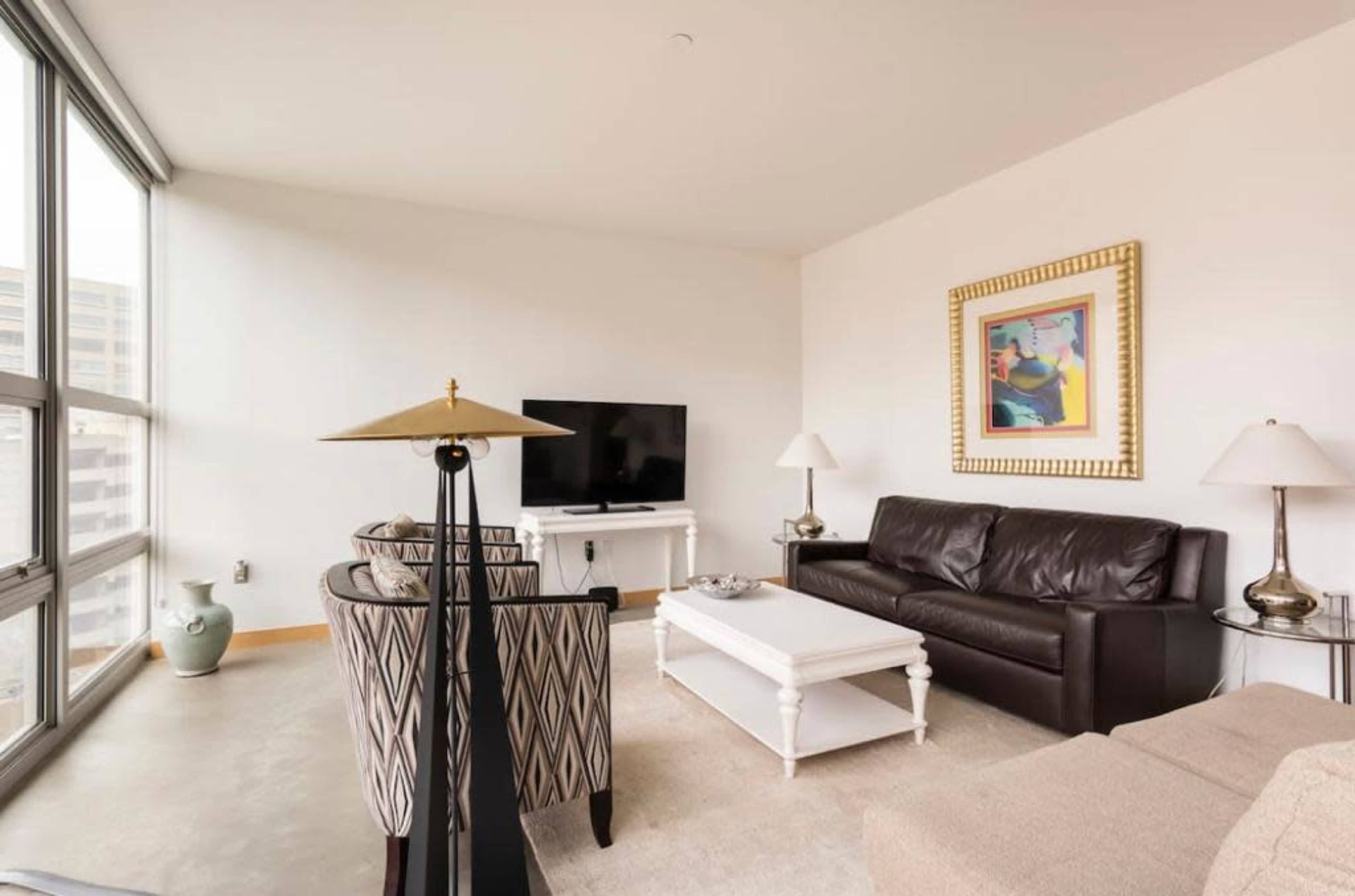 The living room features a leather sofa, two patterned chairs, a coffee table, a television, and large windows providing natural light.