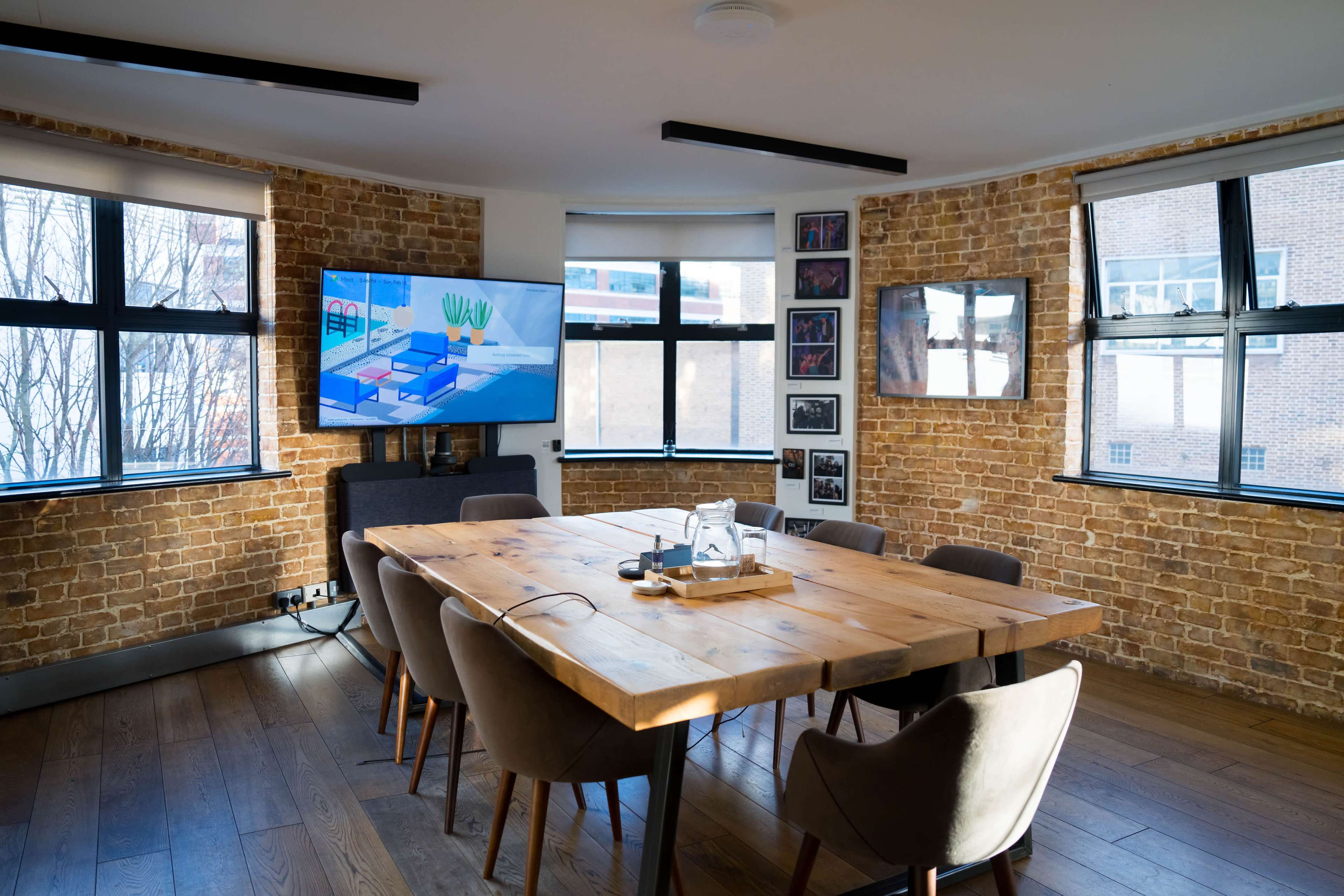 A modern conference room features a large wooden table, comfortable chairs, and a wall-mounted television, with exposed brick walls and multiple windows allowing natural light.