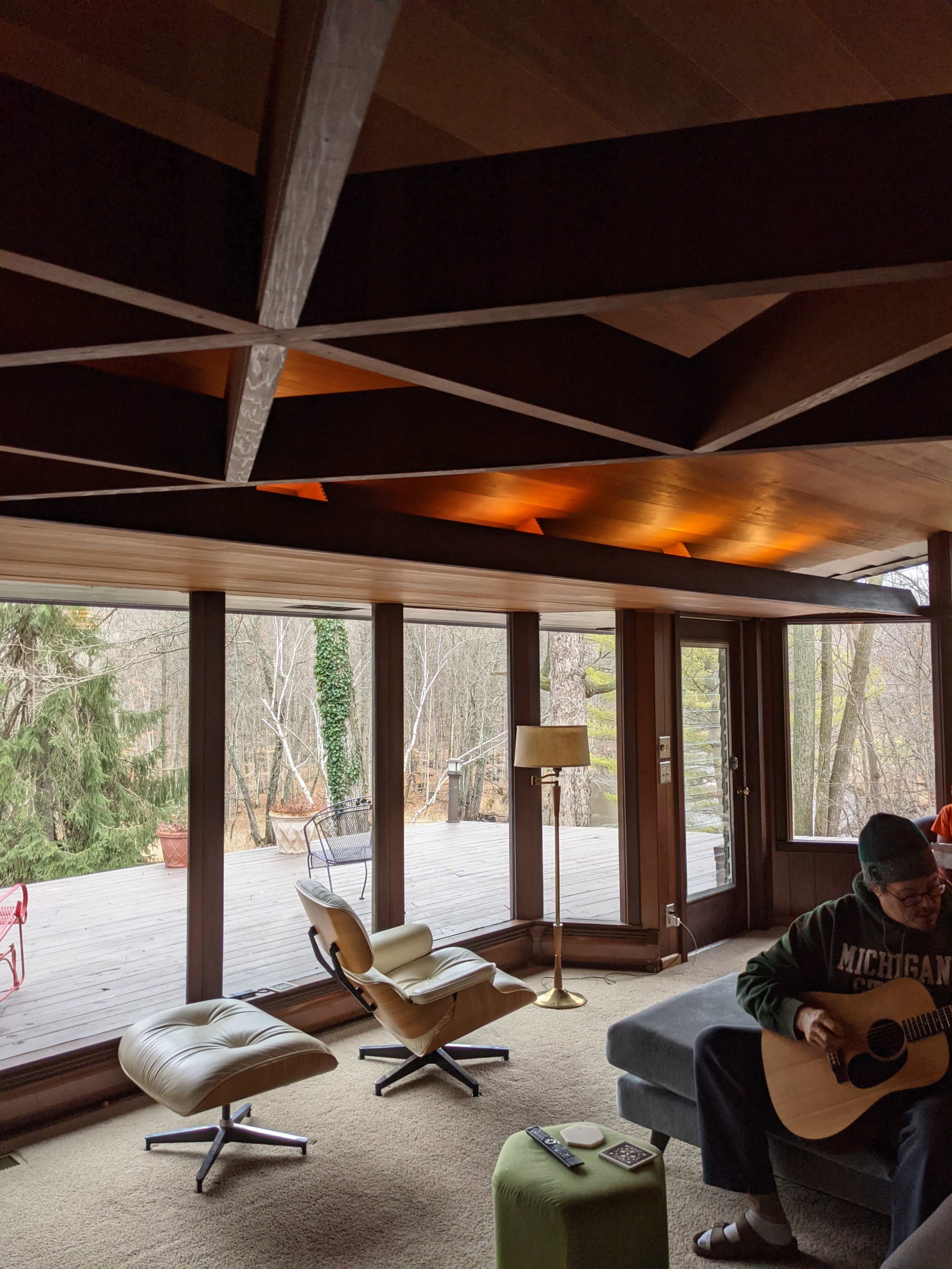 A man sits on a dark sofa playing a guitar in a room with large windows overlooking a wooded area.