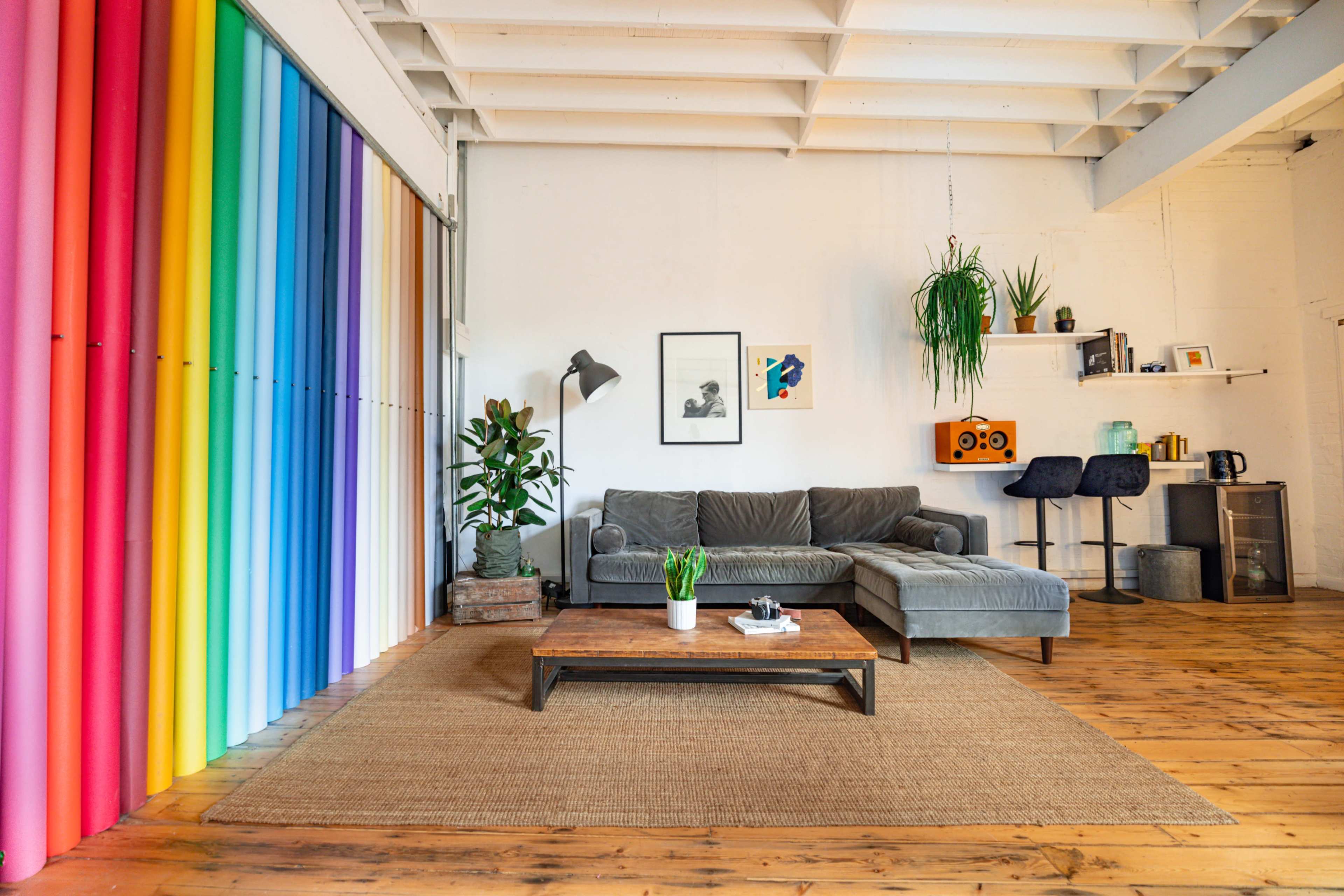 A living room featuring a gray sectional sofa, a colorful wall of vertical blinds, a wooden coffee table, and decorative plants, with a small bar area in the background.