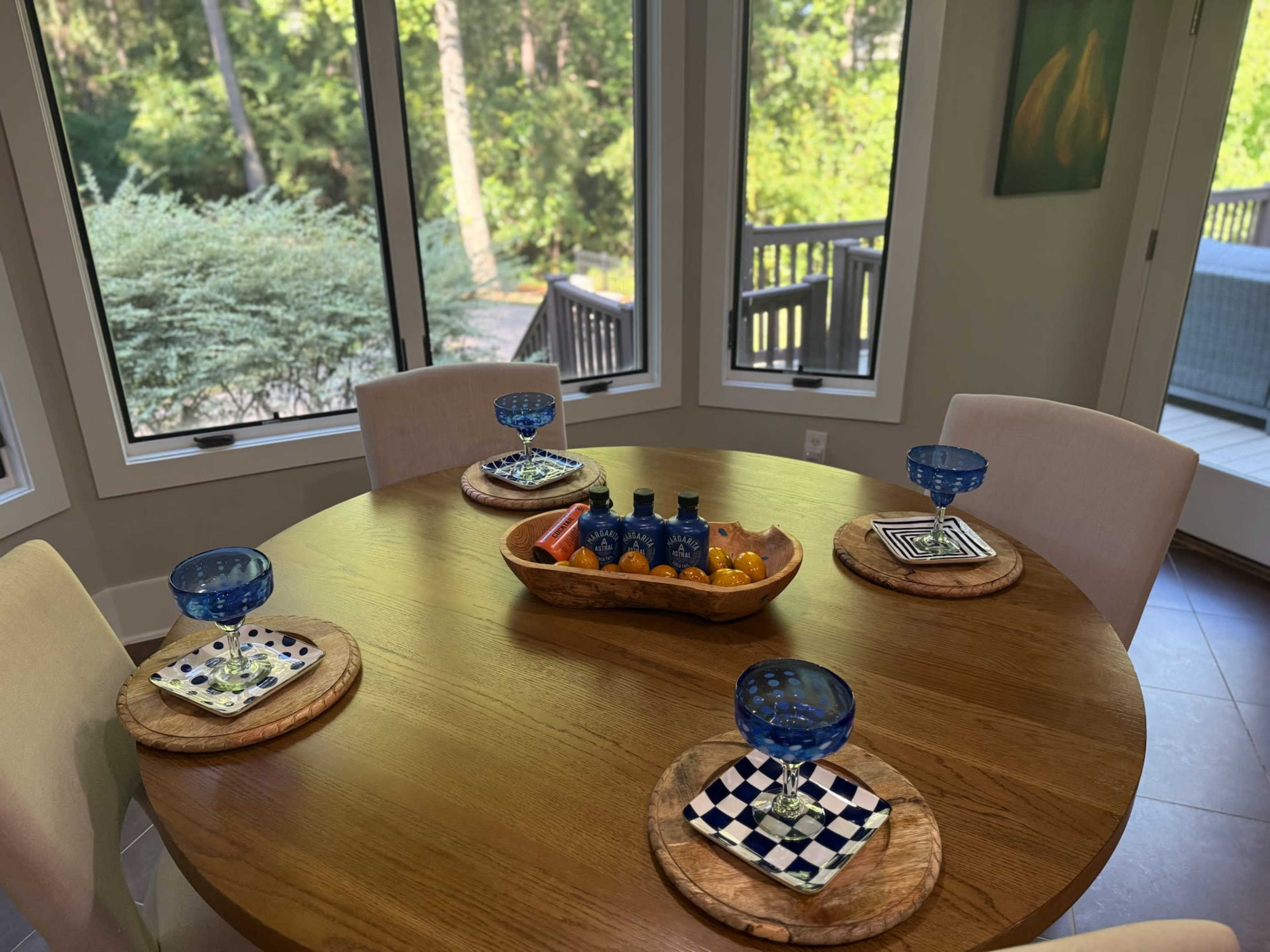 A wooden dining table is set with blue dishes and a centerpiece bowl of fruit, surrounded by light-colored chairs in a sunlit room with large windows overlooking a green landscape.