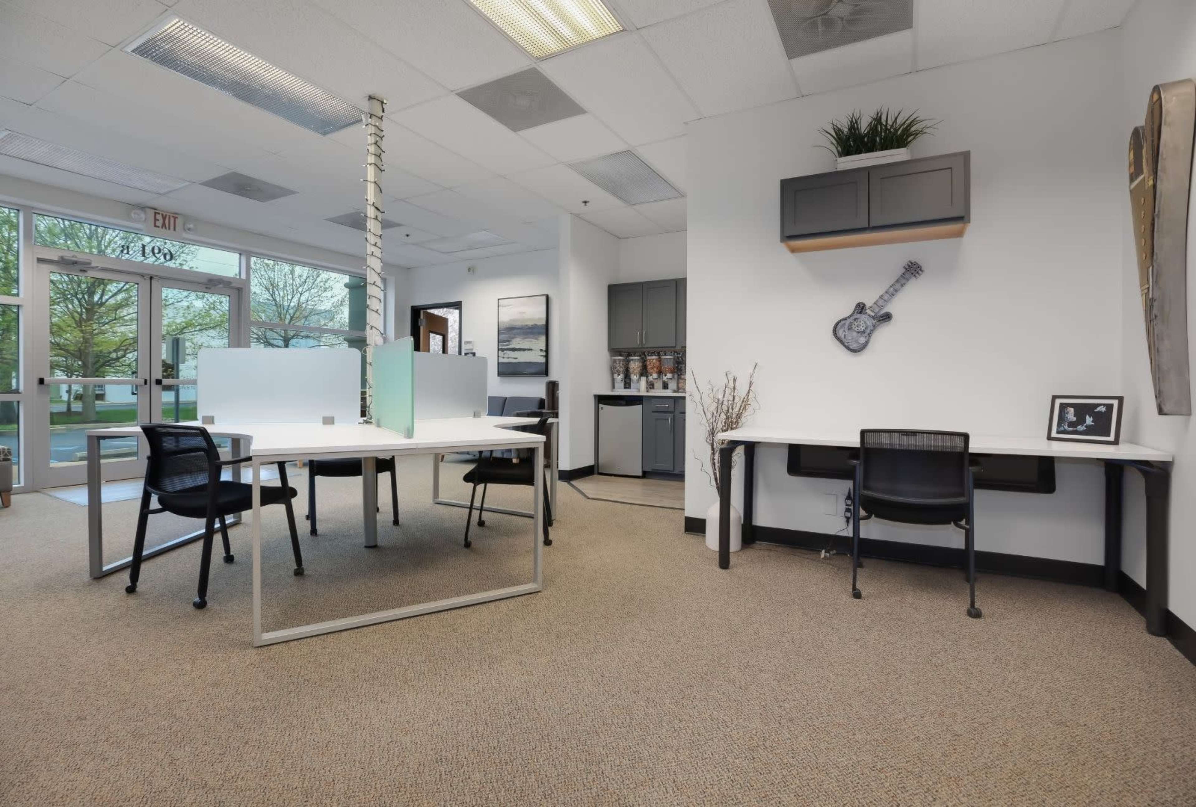 The image shows a modern office space with multiple desks, black chairs, and light-colored walls, featuring a kitchenette and decorative elements.