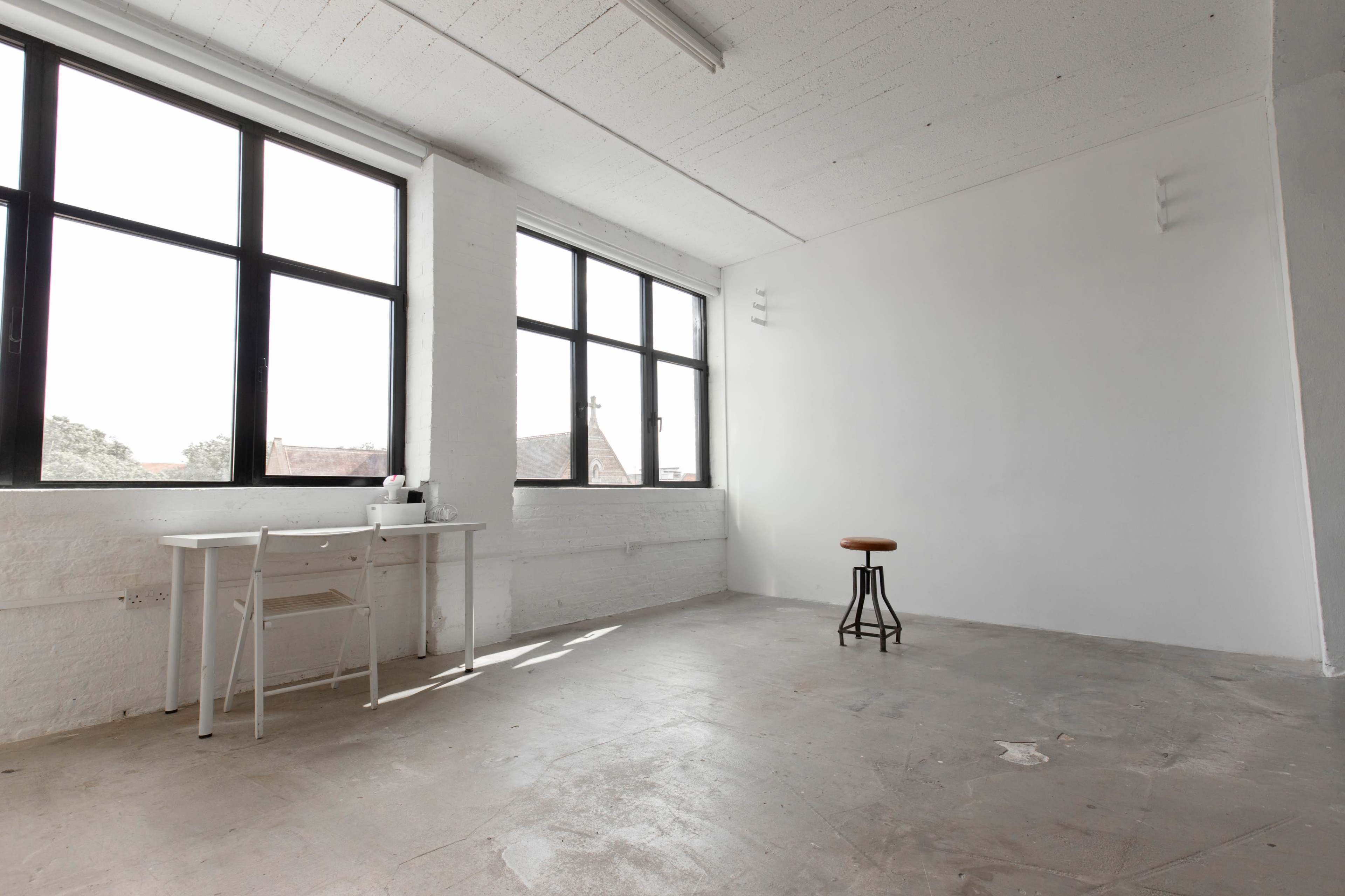 A spacious, empty room with large windows, a small desk on one side, and a stool in the center.