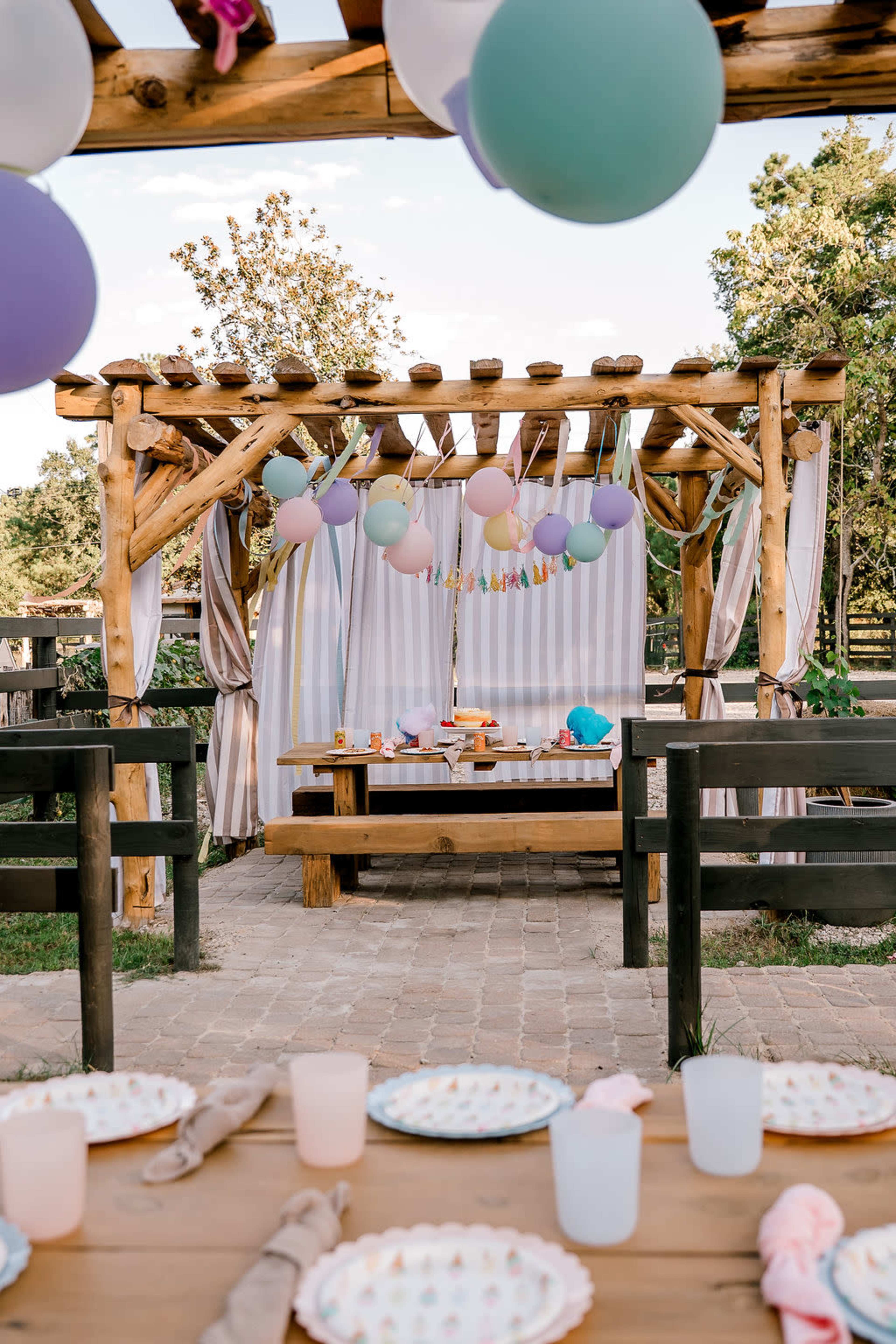 A decorated outdoor seating area with a wooden pergola, colorful balloons hanging above a table set for a party.