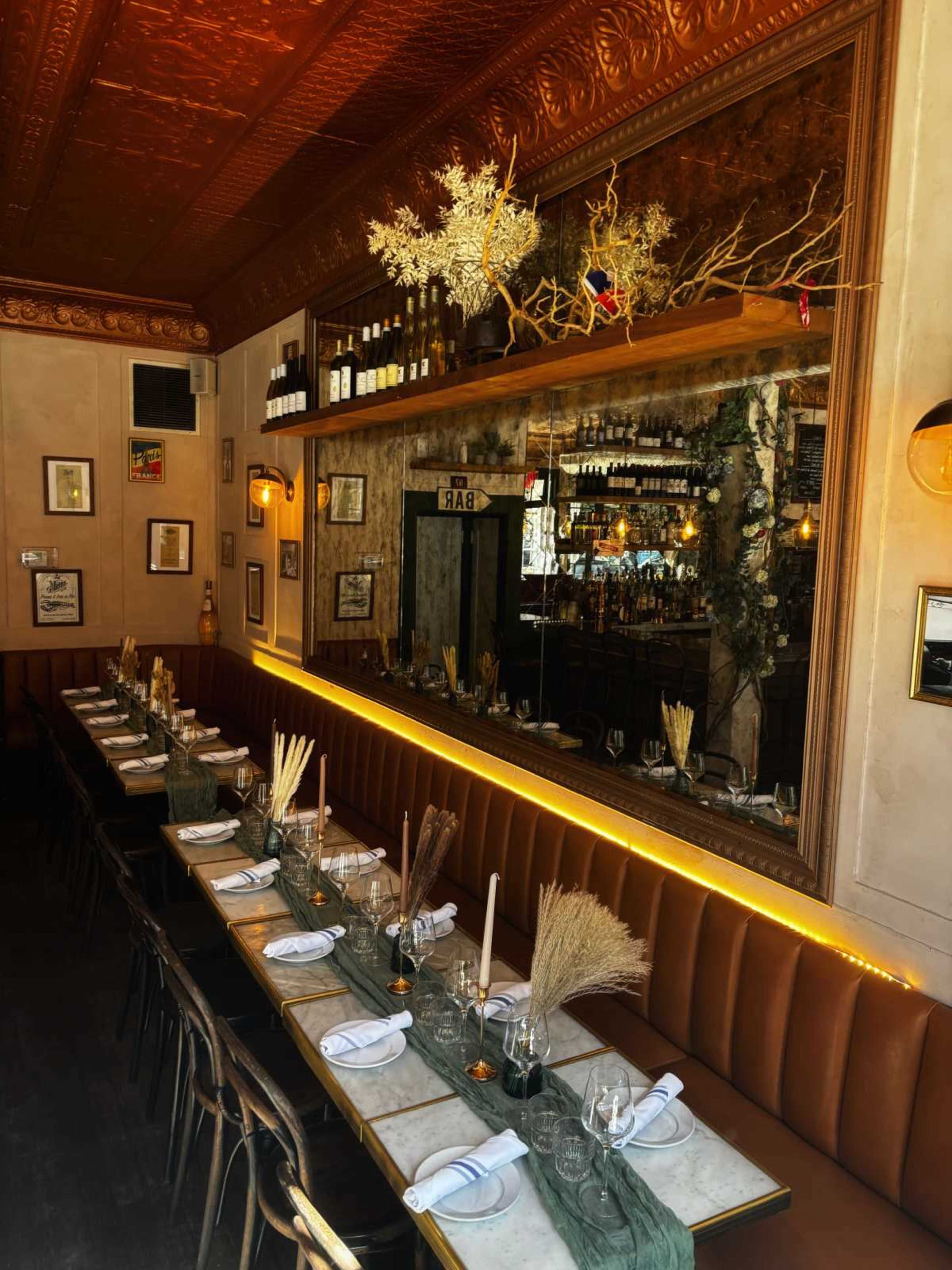 A long dining table is set with white plates, glasses, and decorative elements, against a backdrop of mirrors and shelves filled with bottles in a warmly lit restaurant.