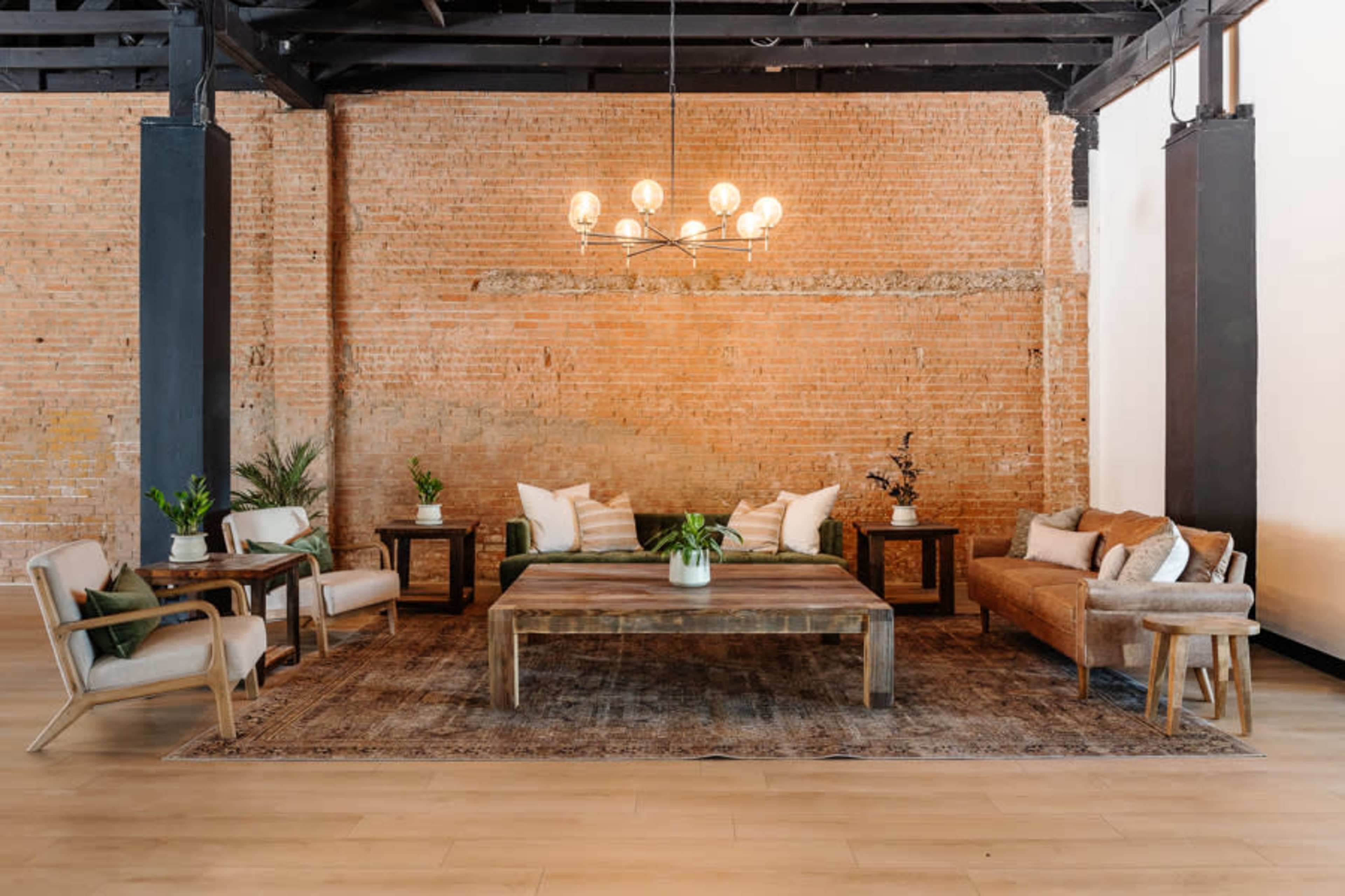 A modern lounge area with a wooden coffee table, various seating arrangements, and potted plants against a brick wall backdrop.
