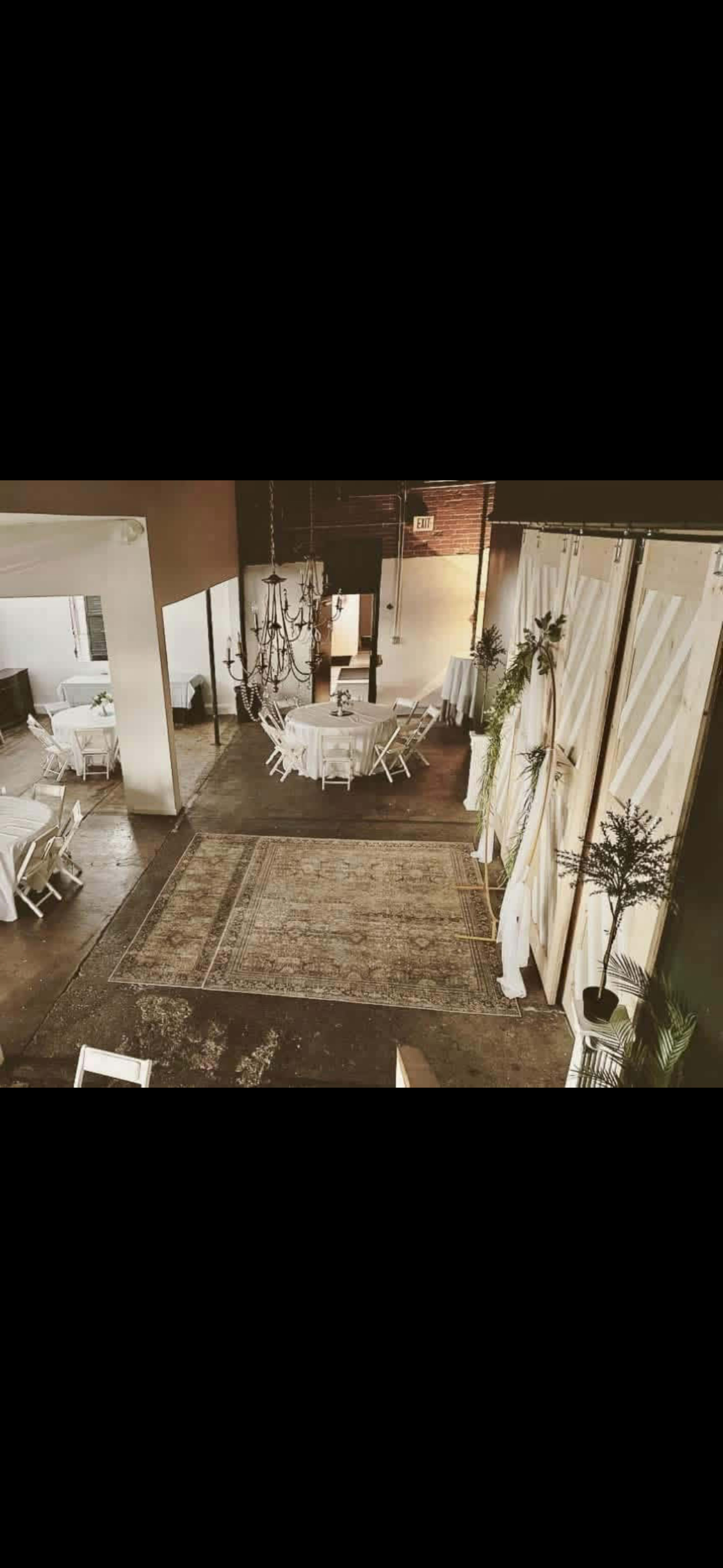 The image shows a spacious venue with several tables set for dining, decorated with white tablecloths and surrounded by greenery, featuring a chandelier and a large area rug on the floor.