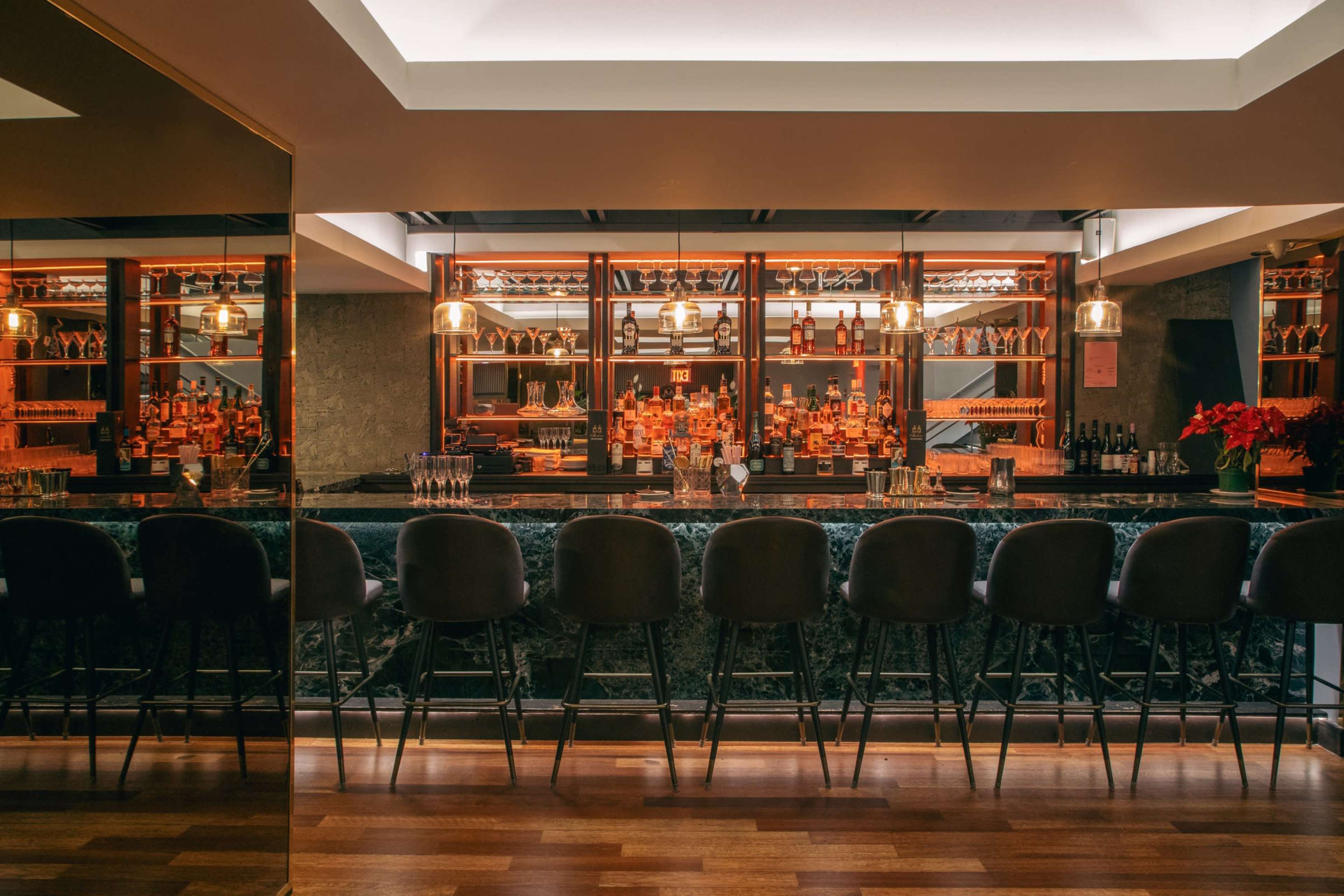 The image shows a modern bar with a polished counter and shelves lined with various bottles behind it, illuminated by pendant lights.
