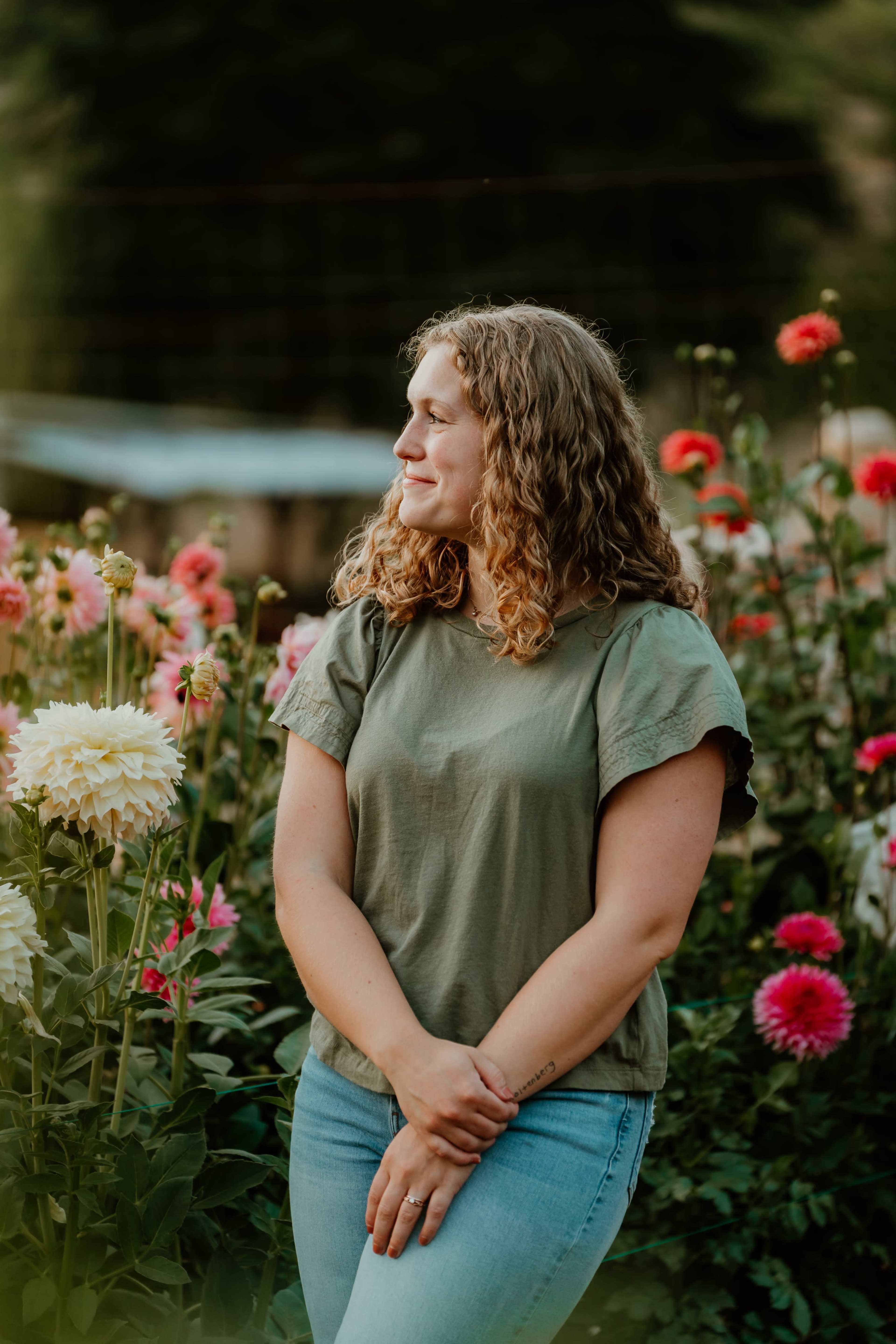 A person stands among vibrant flowers, wearing a green shirt and blue jeans, with a serene expression.