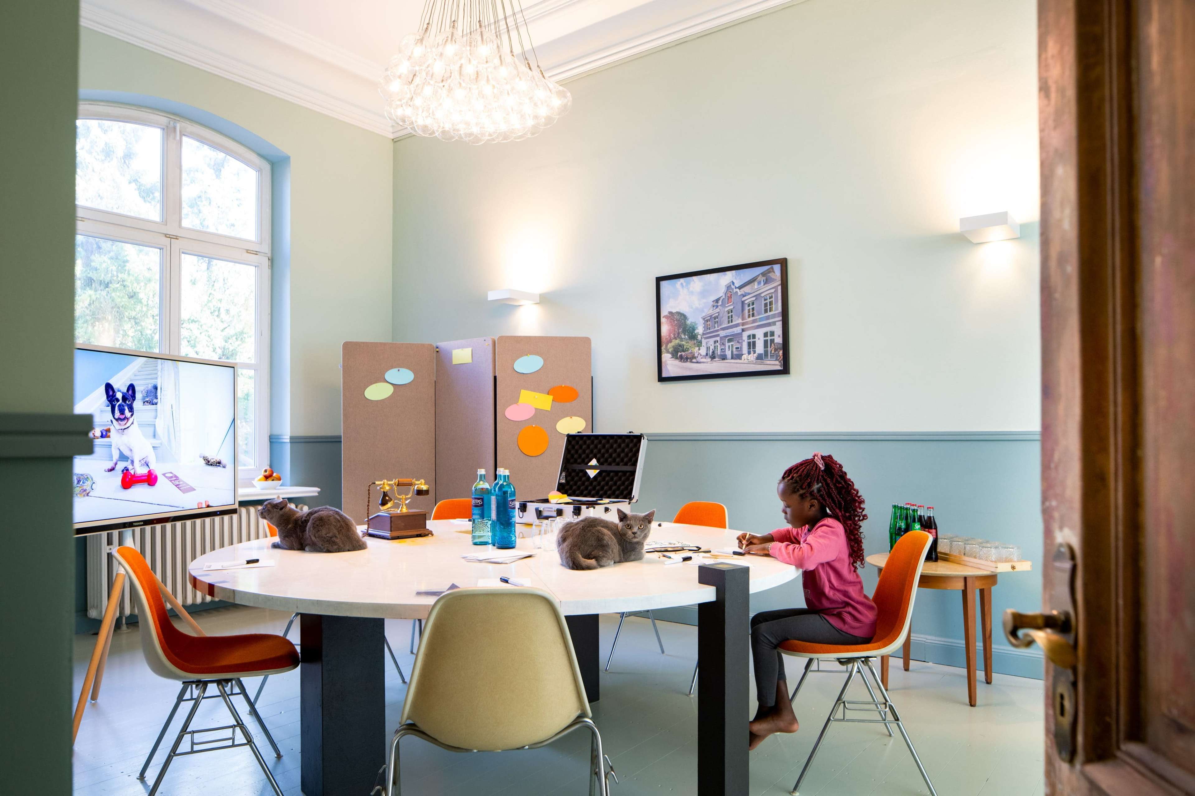 A young girl sits at a round table in a room with two cats, alongside colorful sticky notes and a television displaying images.