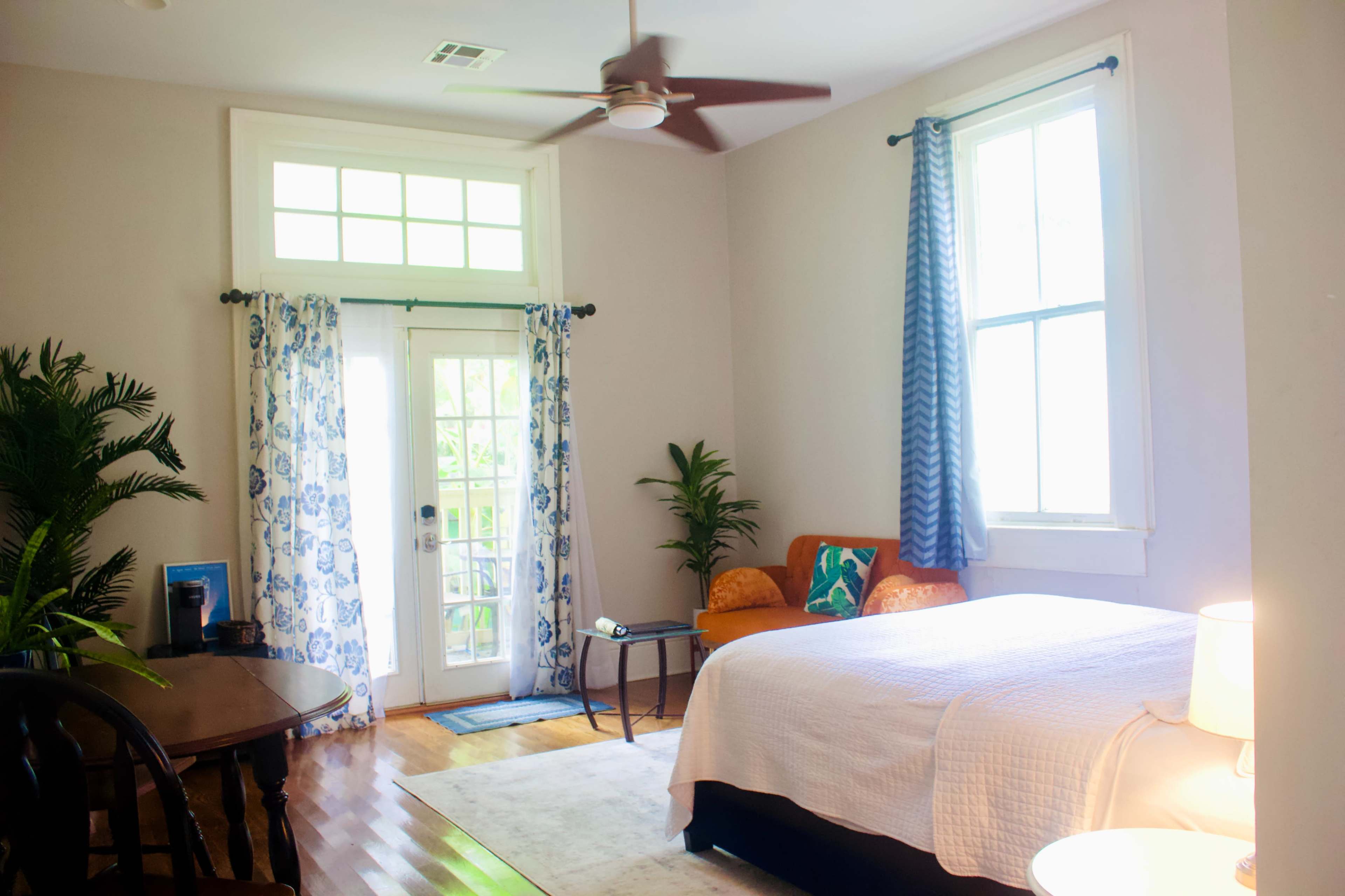 The image shows a bright bedroom featuring a bed, a small table, and a door leading to a patio, with light-colored walls and blue curtains.