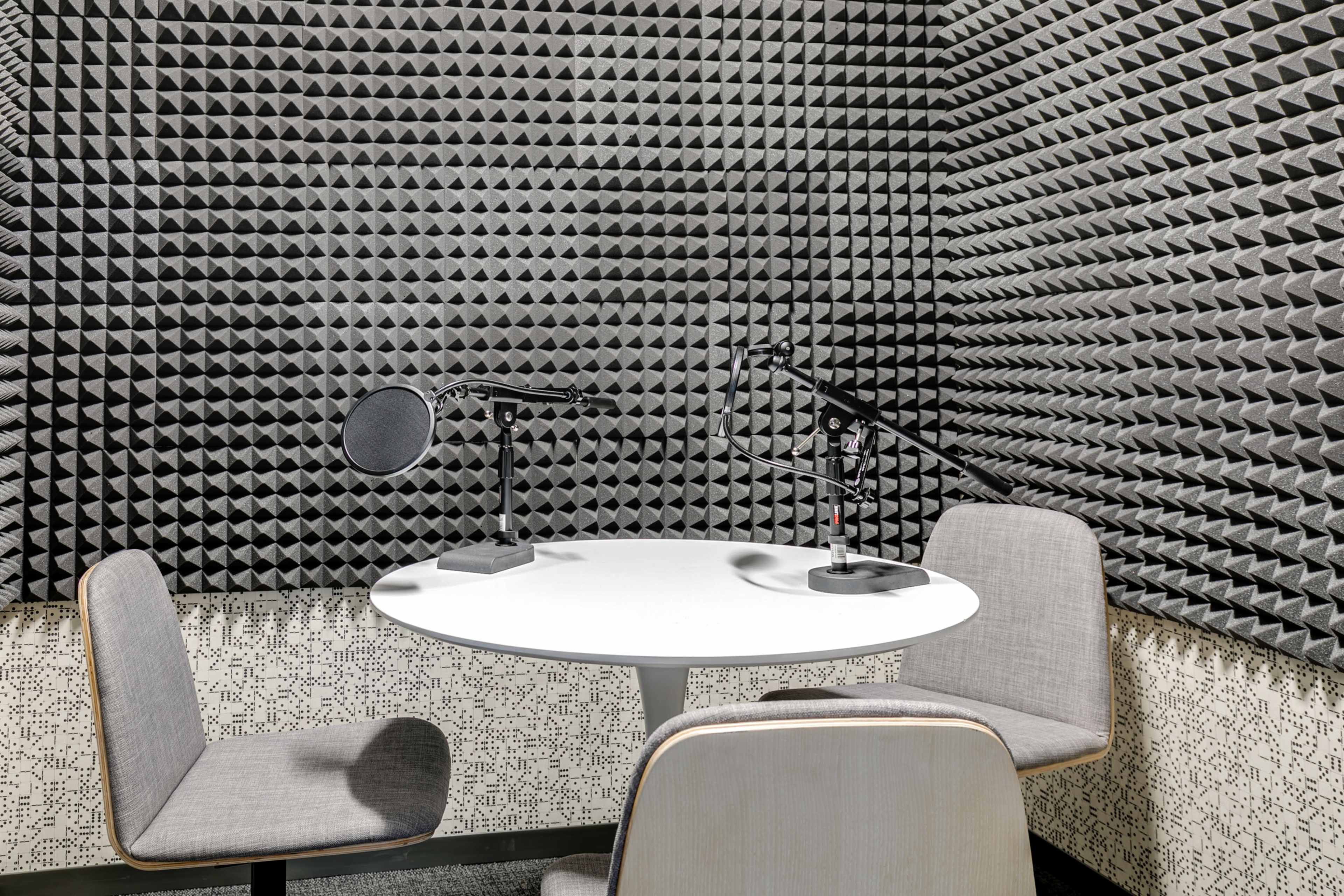 The image shows a small recording studio with soundproof walls, a round table, and two chairs, each equipped with a microphone on stands.
