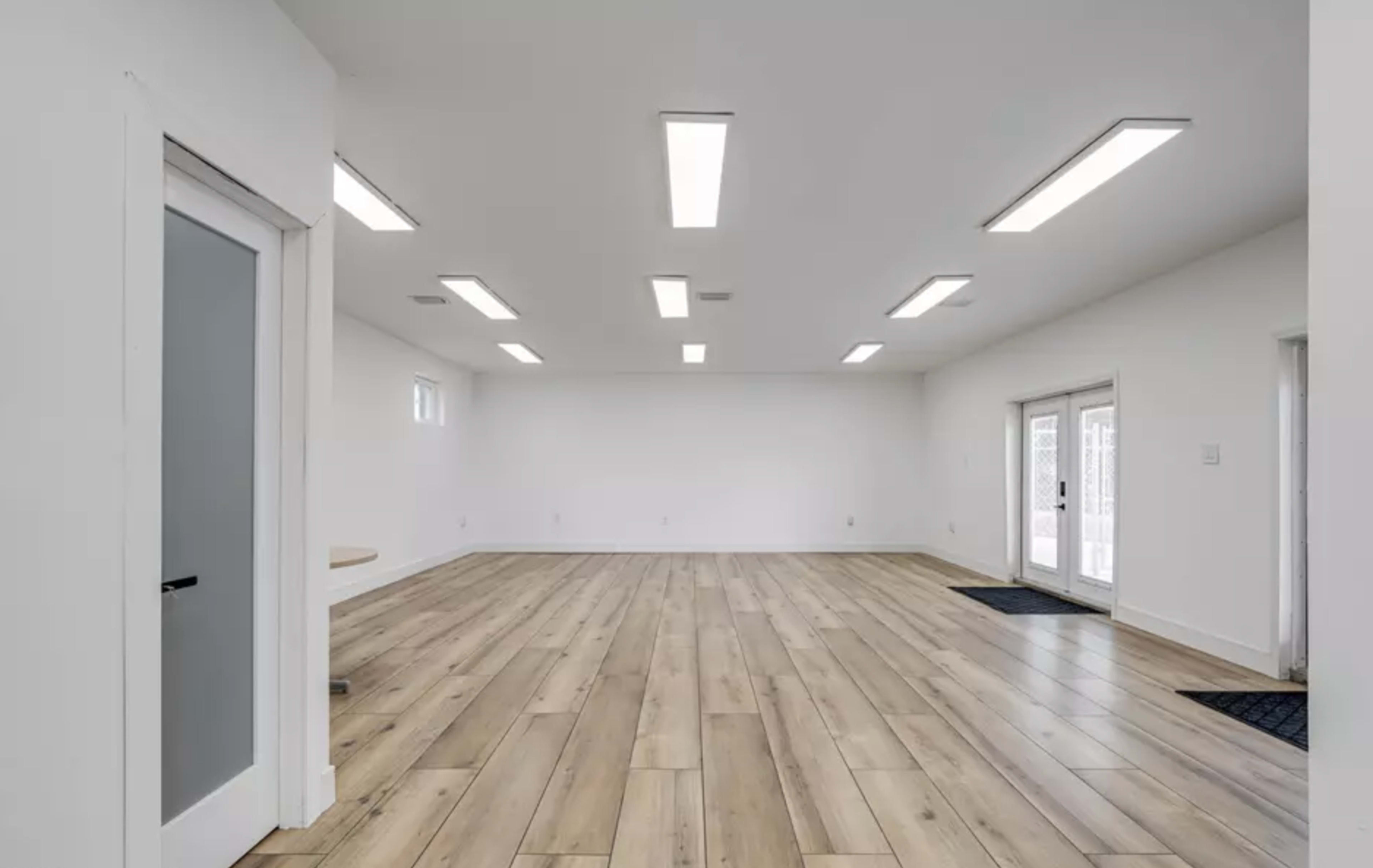 The image shows a large, empty room with wooden flooring and white walls, featuring multiple fluorescent lights overhead.