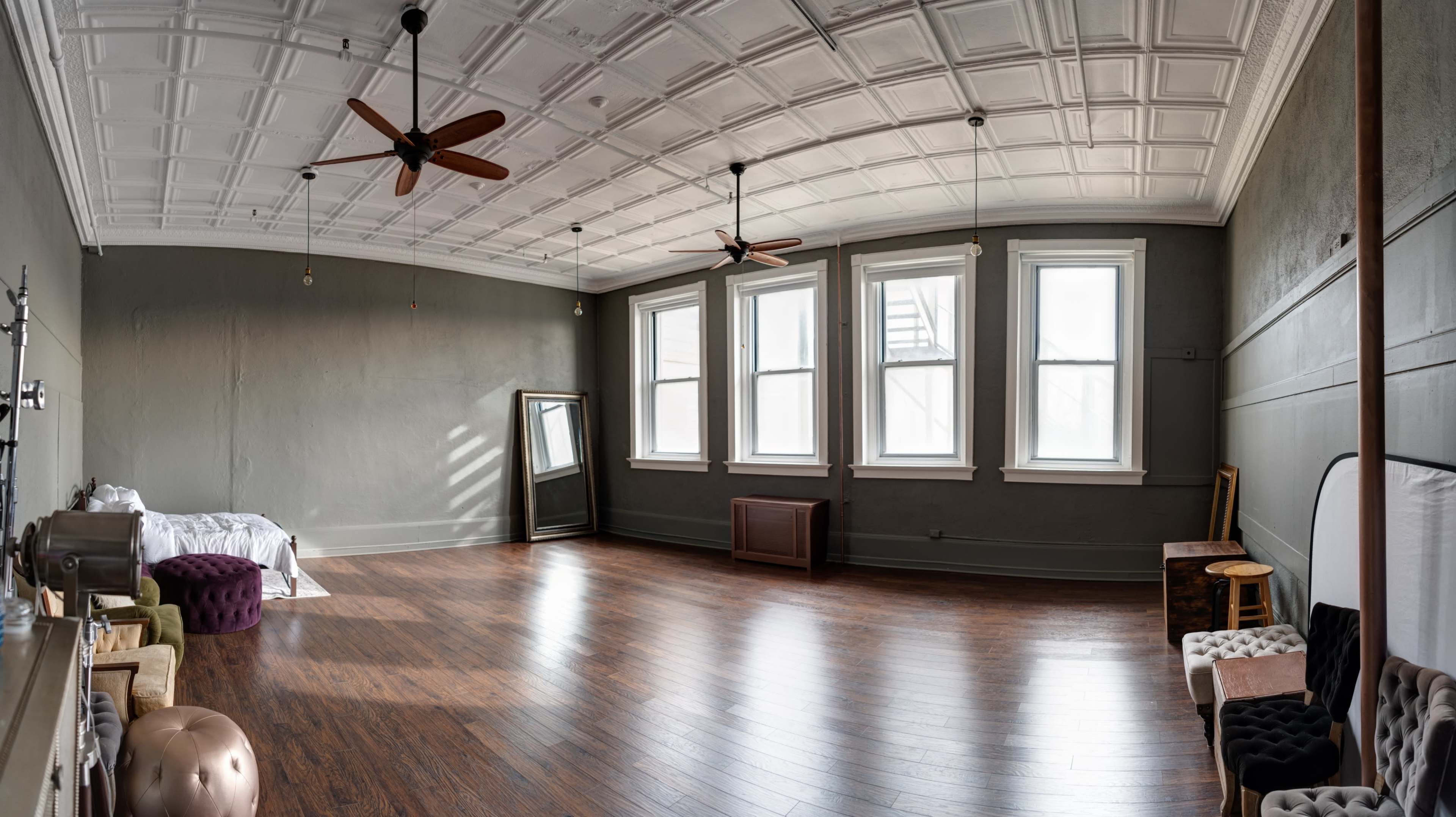 A spacious room with a wooden floor, large windows, a mirror, and ceiling fans, featuring a minimalistic design.