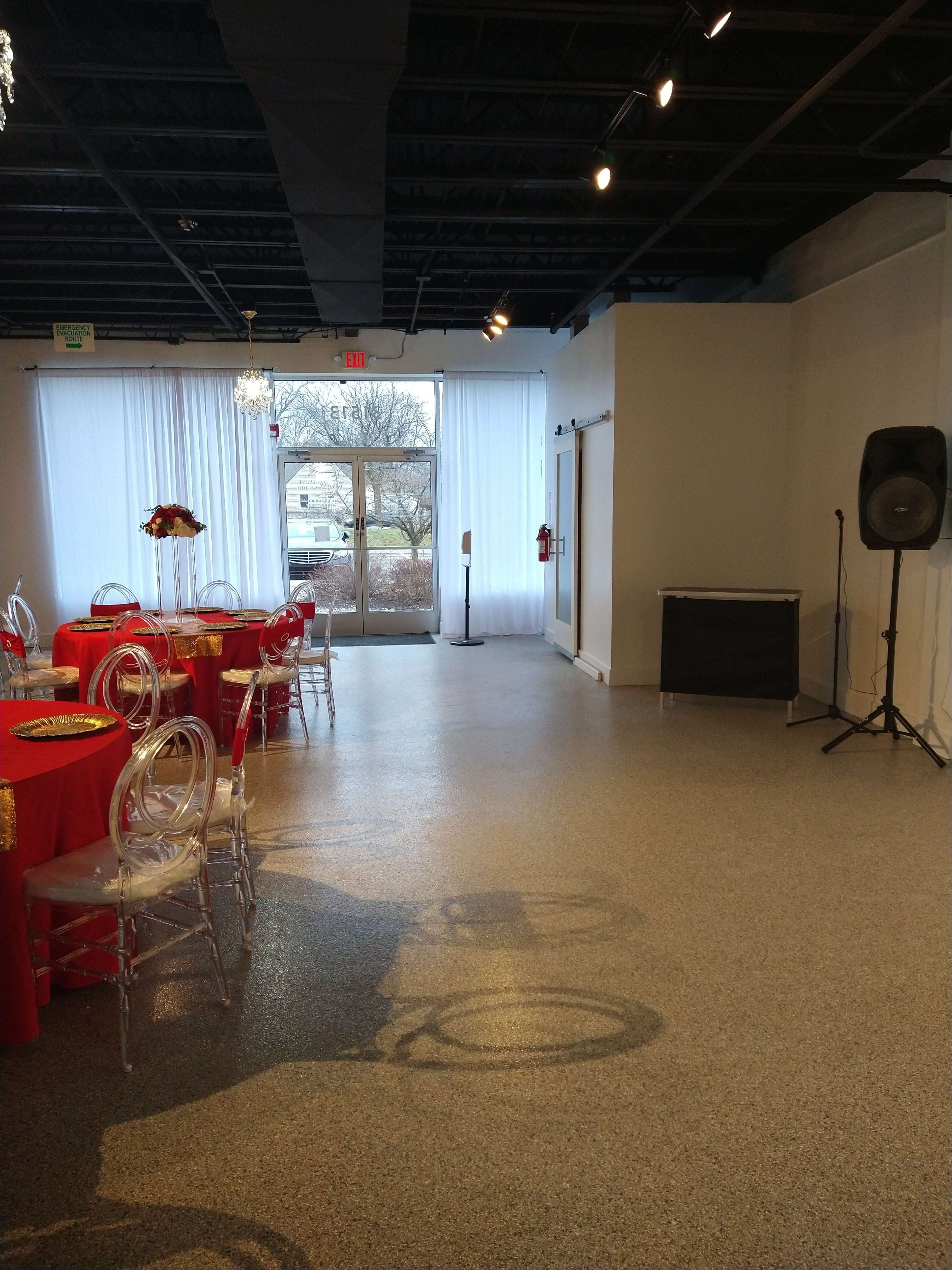 The image shows an empty event space with red-covered tables, clear chairs, and large windows letting in natural light.