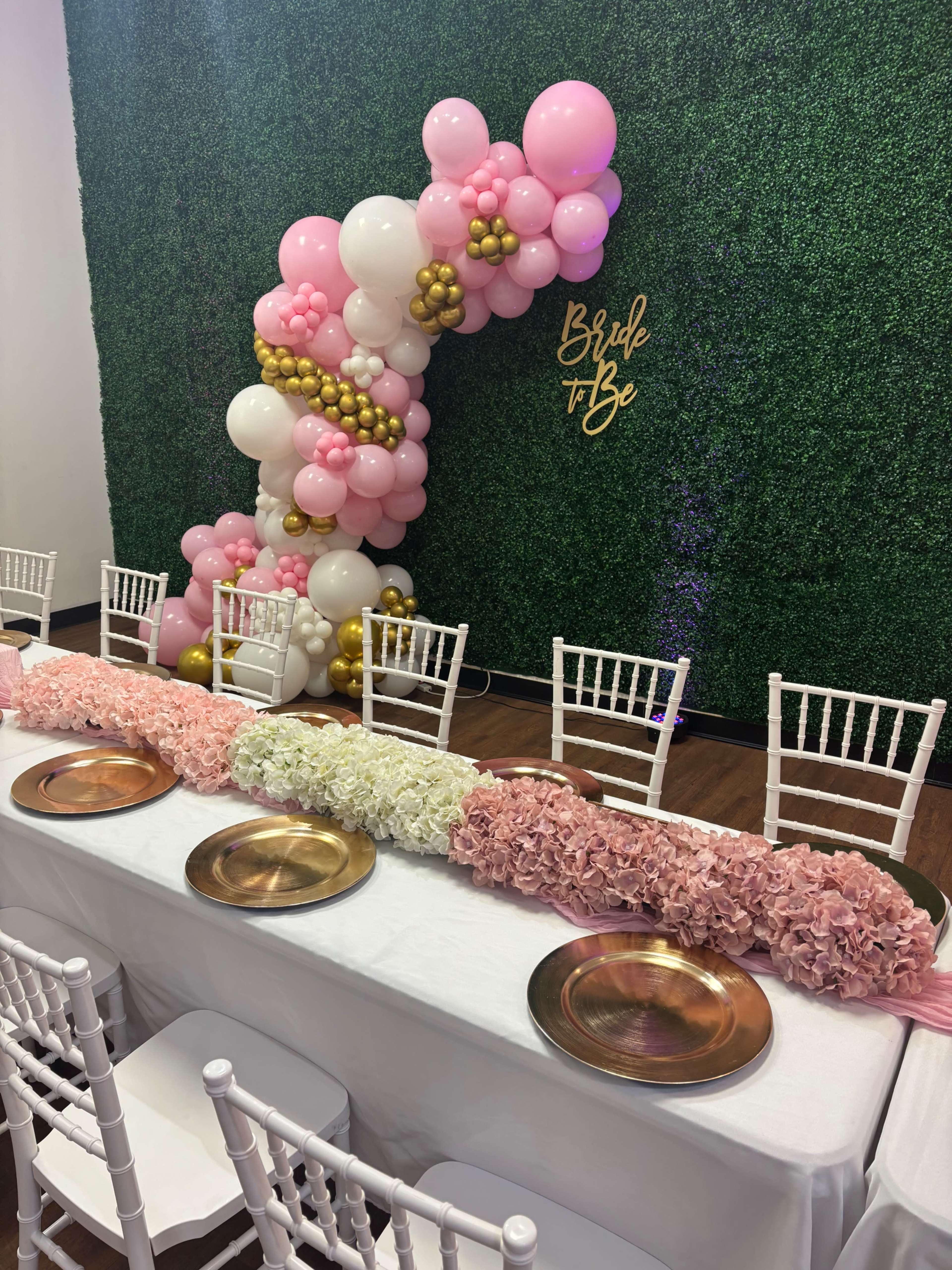 A decorated table with a floral centerpiece is set against a green foliage backdrop and an arch made of balloons in pink, white, and gold.