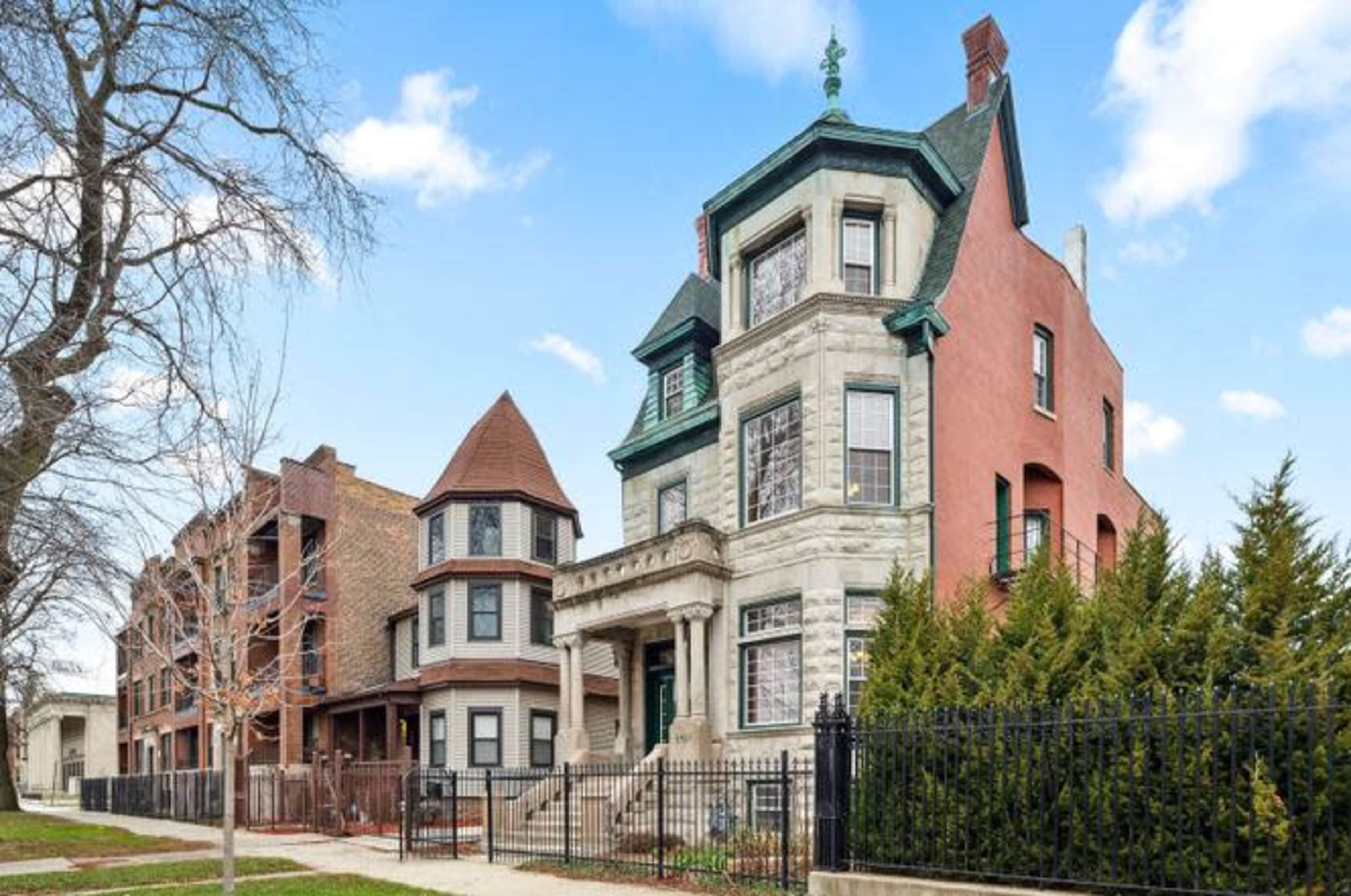 The image shows a historic multi-story brick building with a distinct architectural design, flanked by other residential structures and greenery along the sidewalk.