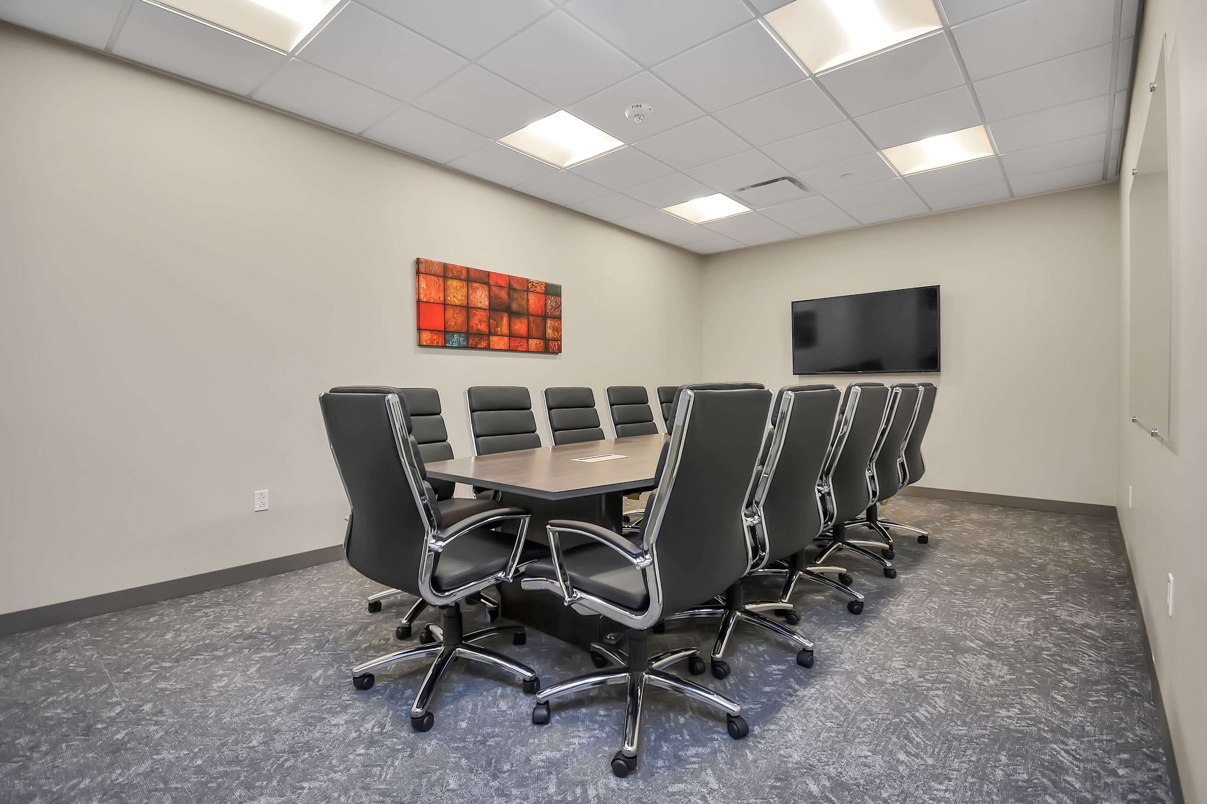 A conference room features a long table surrounded by black ergonomic chairs, with a wall-mounted TV and artwork in the background.