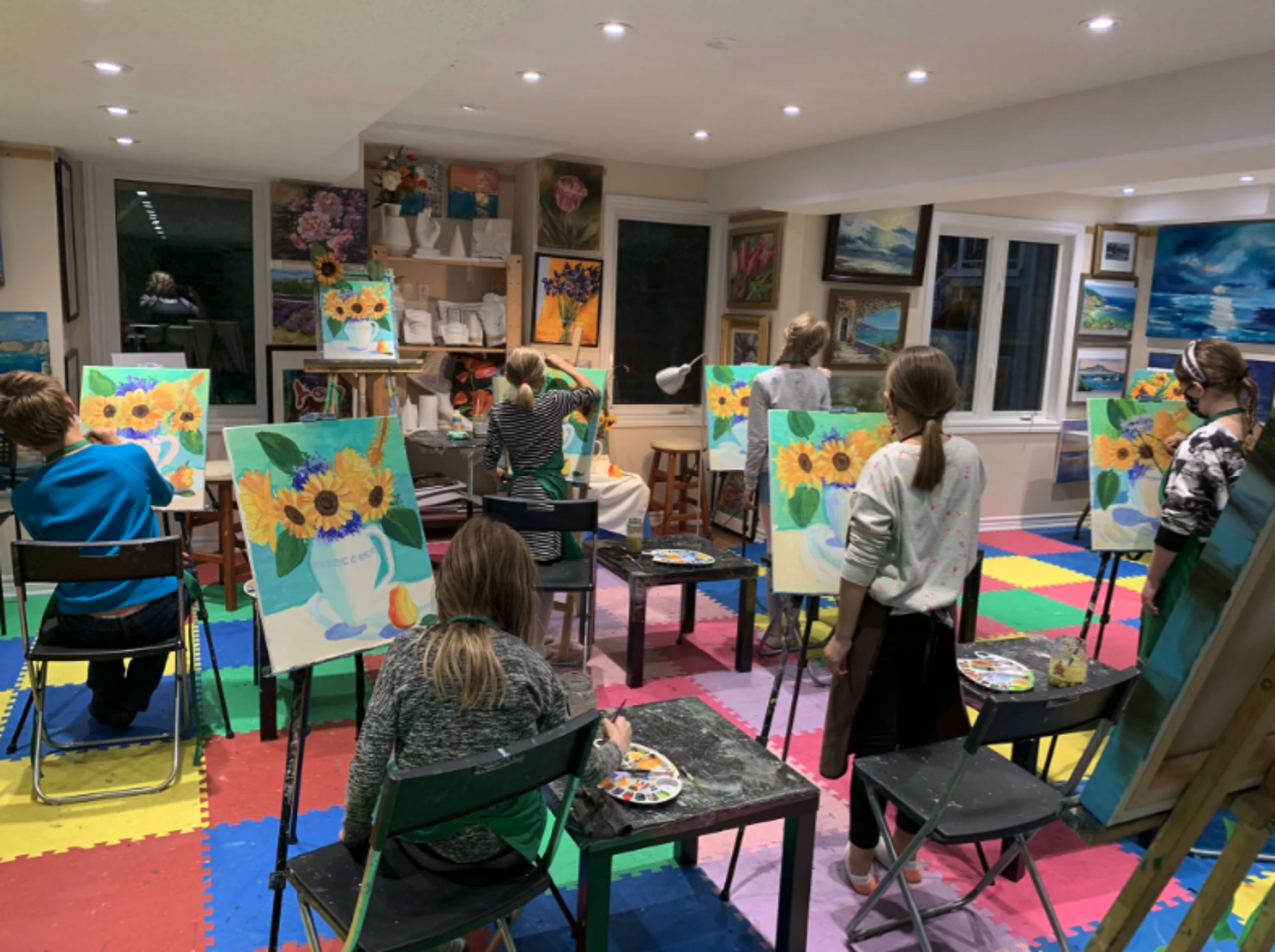 A group of children sits at easels, painting sunflowers in a colorful art studio with various paintings displayed on the walls.