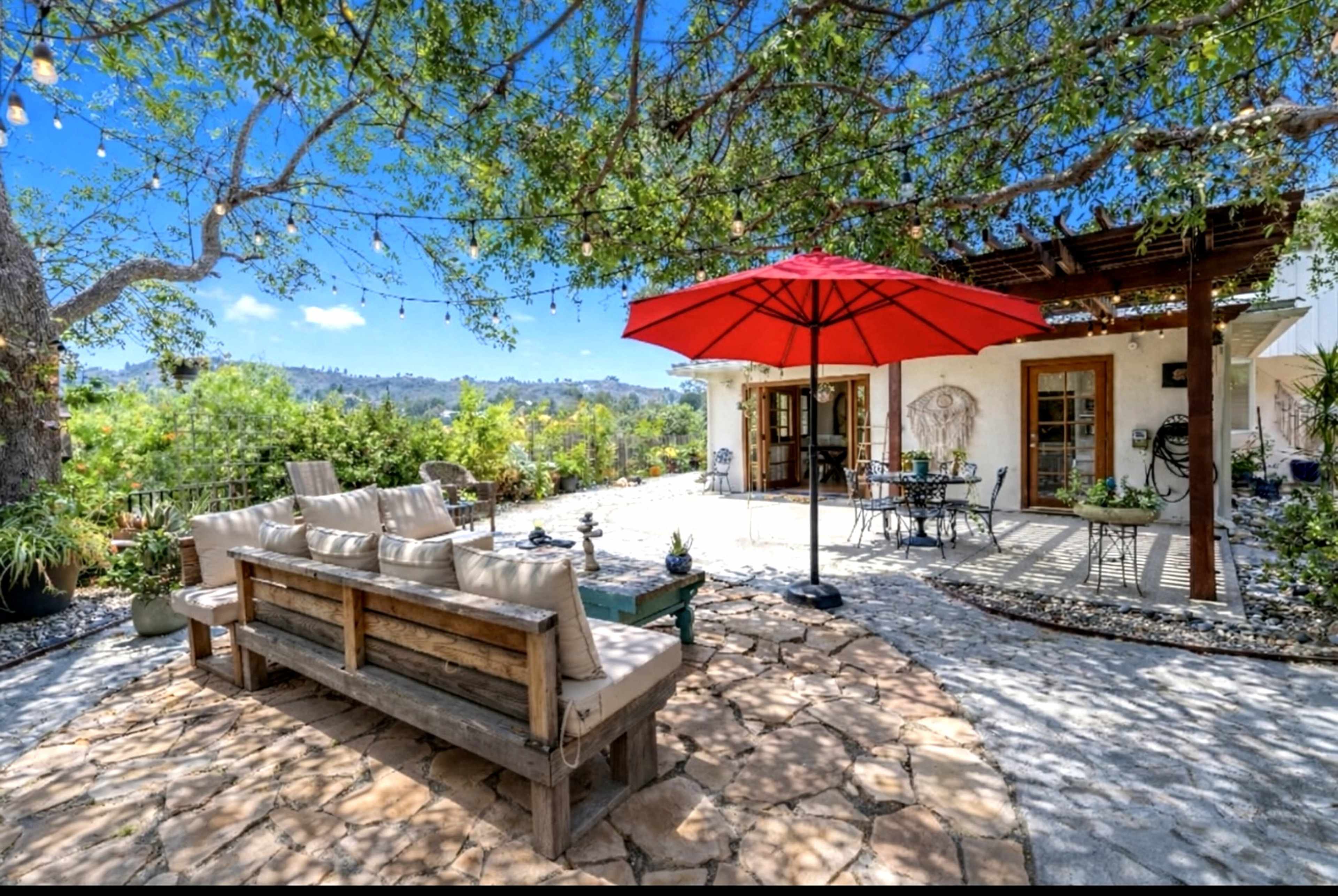 The image shows a spacious outdoor patio area surrounded by greenery, featuring a wooden bench, a red umbrella, and seating arrangements on stone flooring.