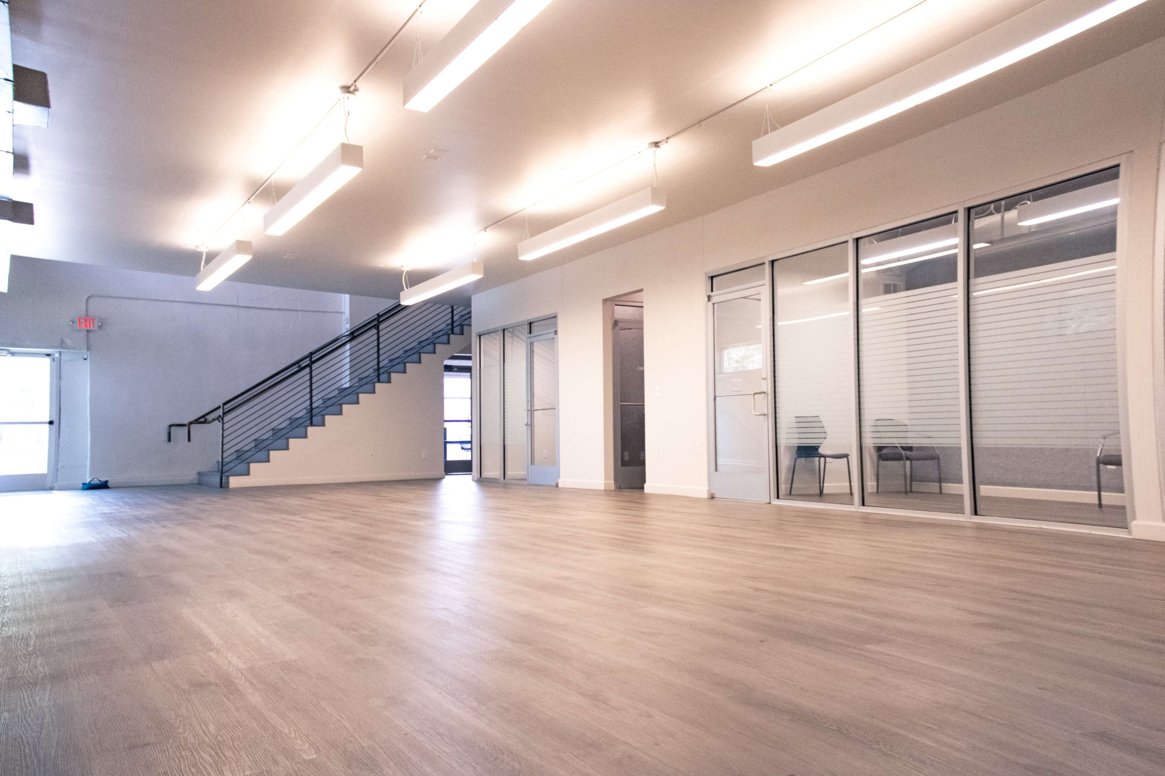 The image shows a spacious, modern interior with large windows, wooden flooring, and a staircase leading to an upper level.