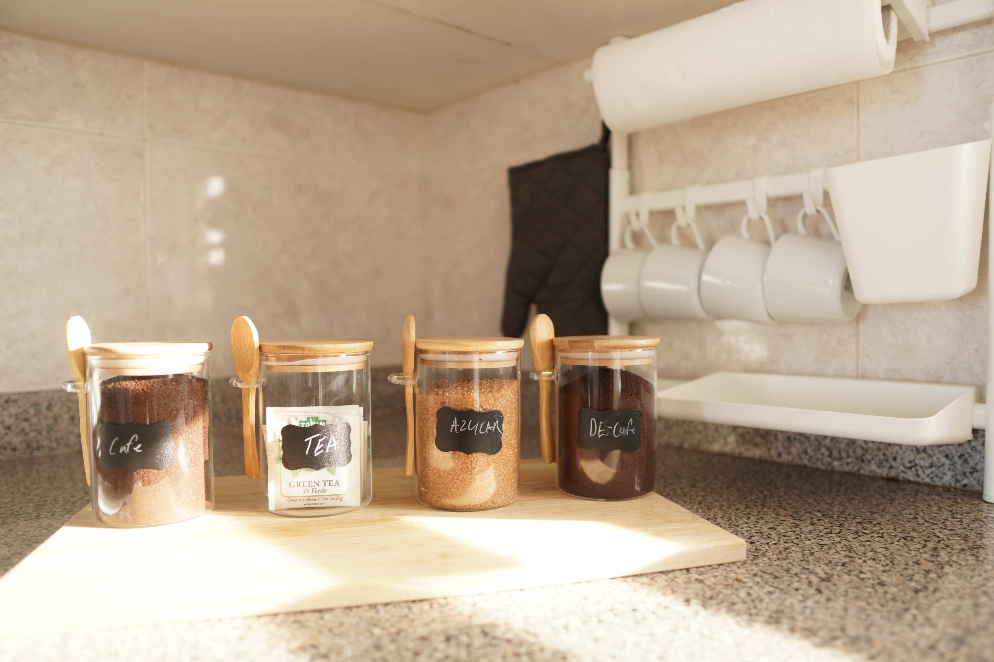 Four glass jars with wooden lids containing labeled contents, including café, tea, azúcar, and decaf, are arranged on a wooden cutting board in a kitchen.
