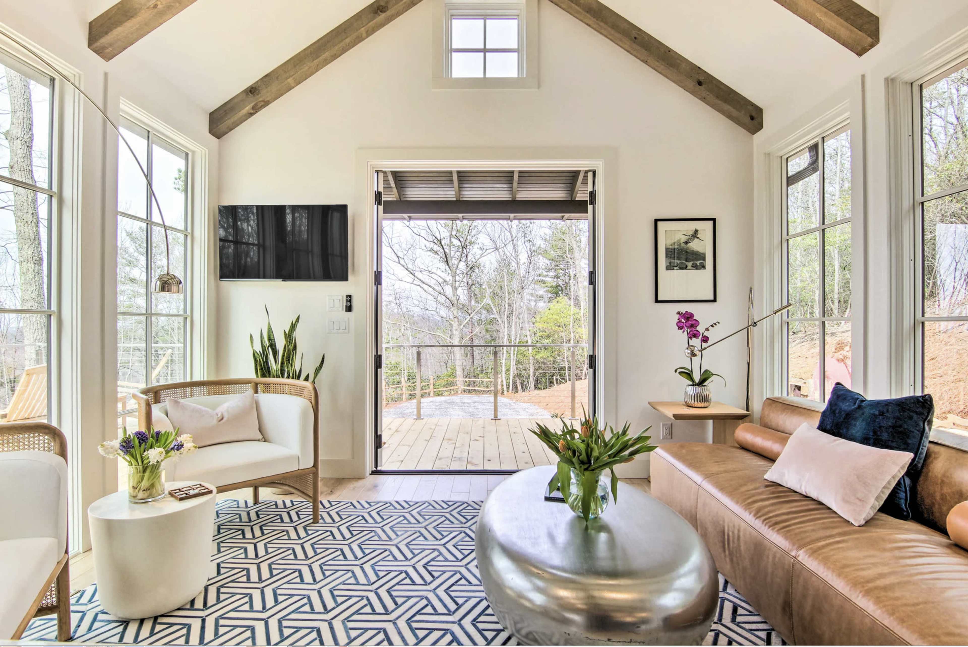 A bright interior features a seating area with a sofa, chairs, and a coffee table, overlooking a deck through large windows.