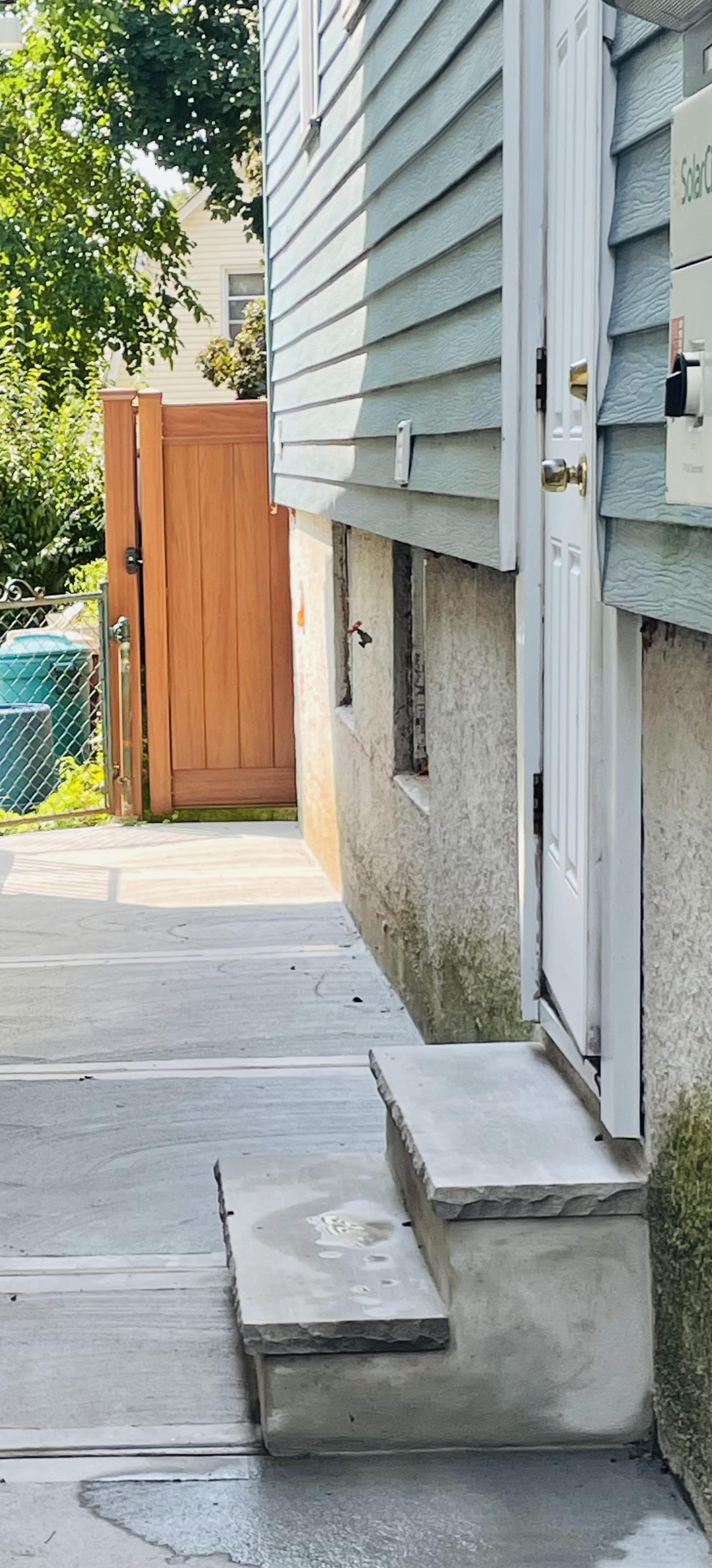 The image shows a concrete pathway leading to a white door at the side of a house, with a wooden gate and a green fence in the background.