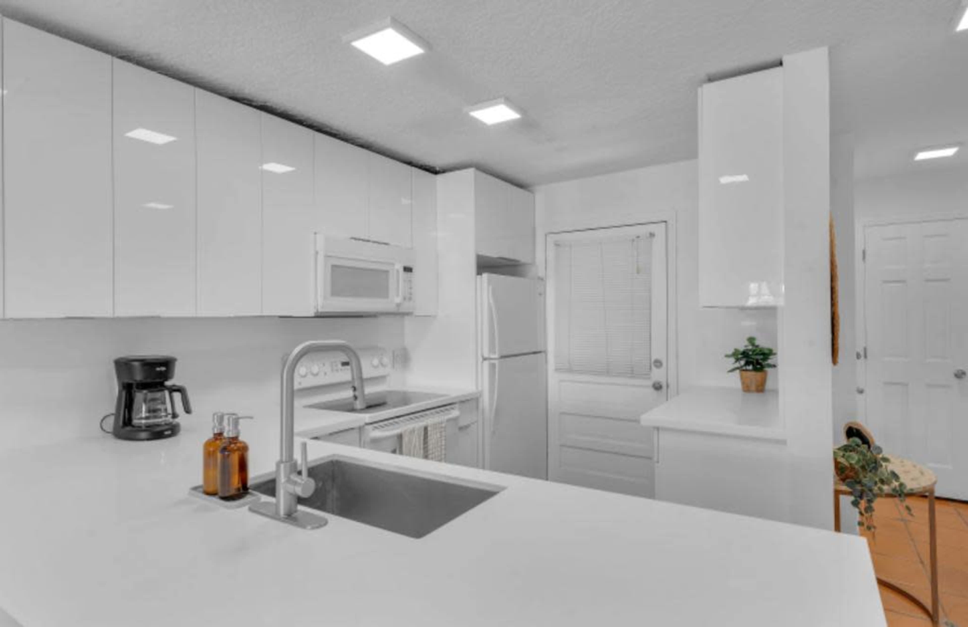A modern kitchen with white cabinets, a stainless steel sink, and an island with a coffee maker and glass bottles on it.