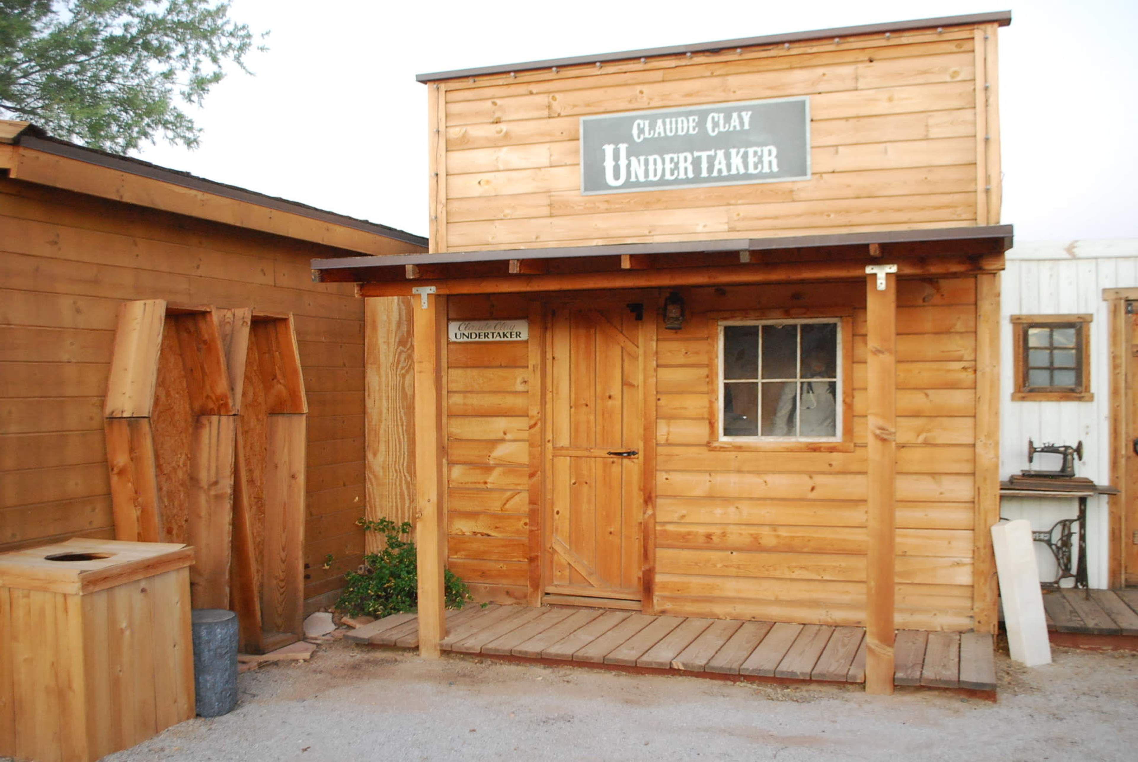 The image shows a wooden building with a sign that reads "Claude Clay Undertaker" above the door, surrounded by other wooden structures.
