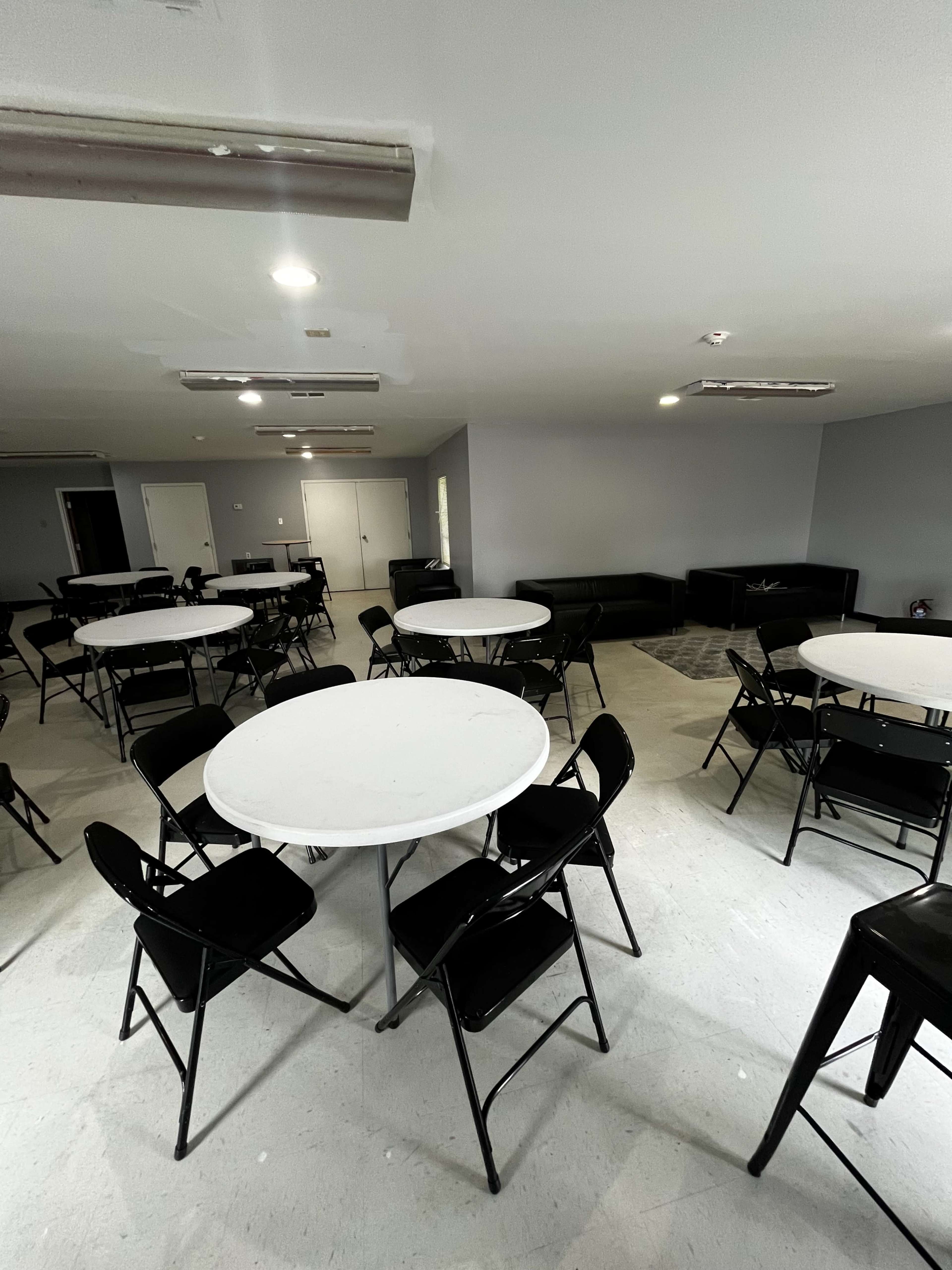 The image shows a spacious room with several circular tables surrounded by black folding chairs, along with a dark couch and gray walls.