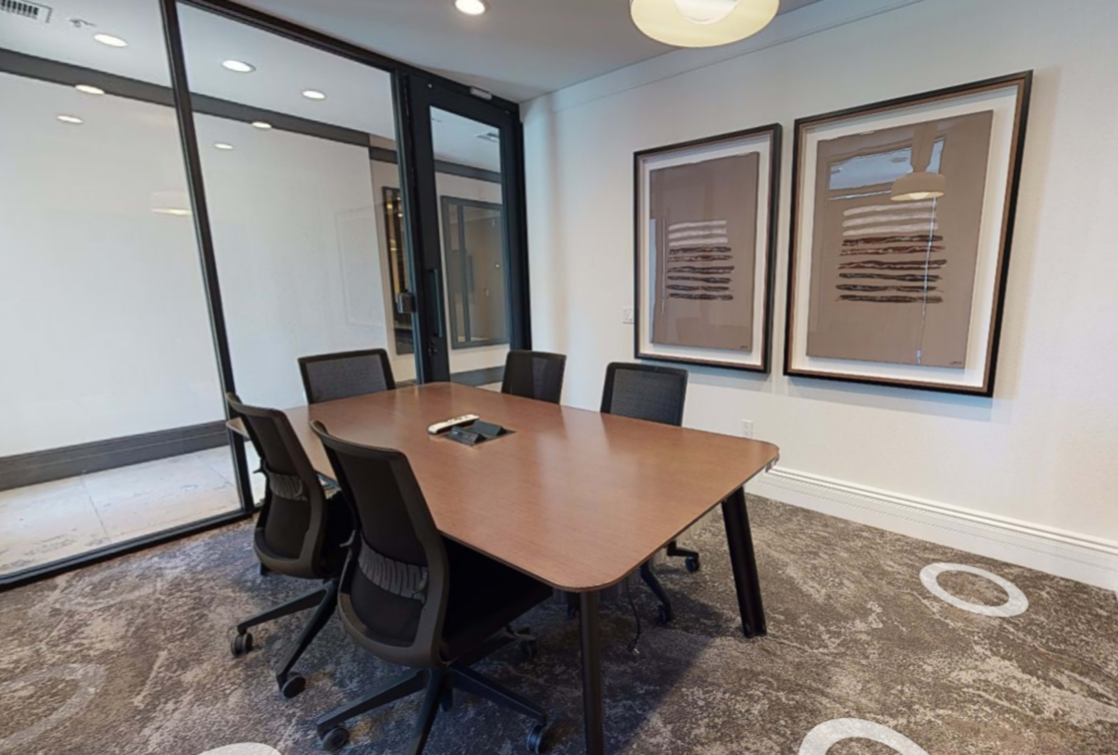 A conference room features a wooden table surrounded by five black chairs, with two framed artworks on the walls and glass walls allowing visibility to the outside.