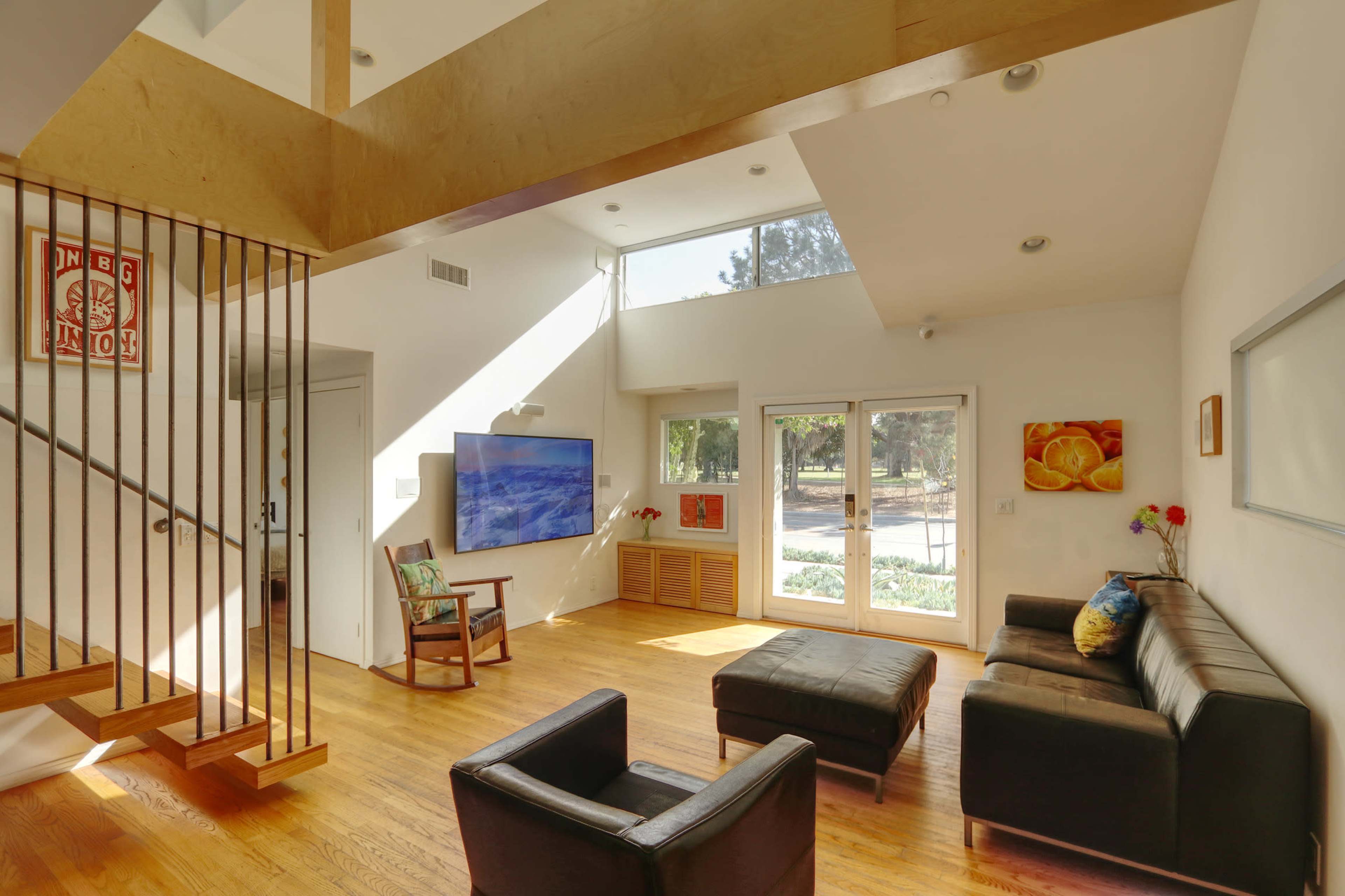 A bright living room with a minimalist design featuring a sectional sofa, two chairs, a television on the wall, and large windows that provide natural light.