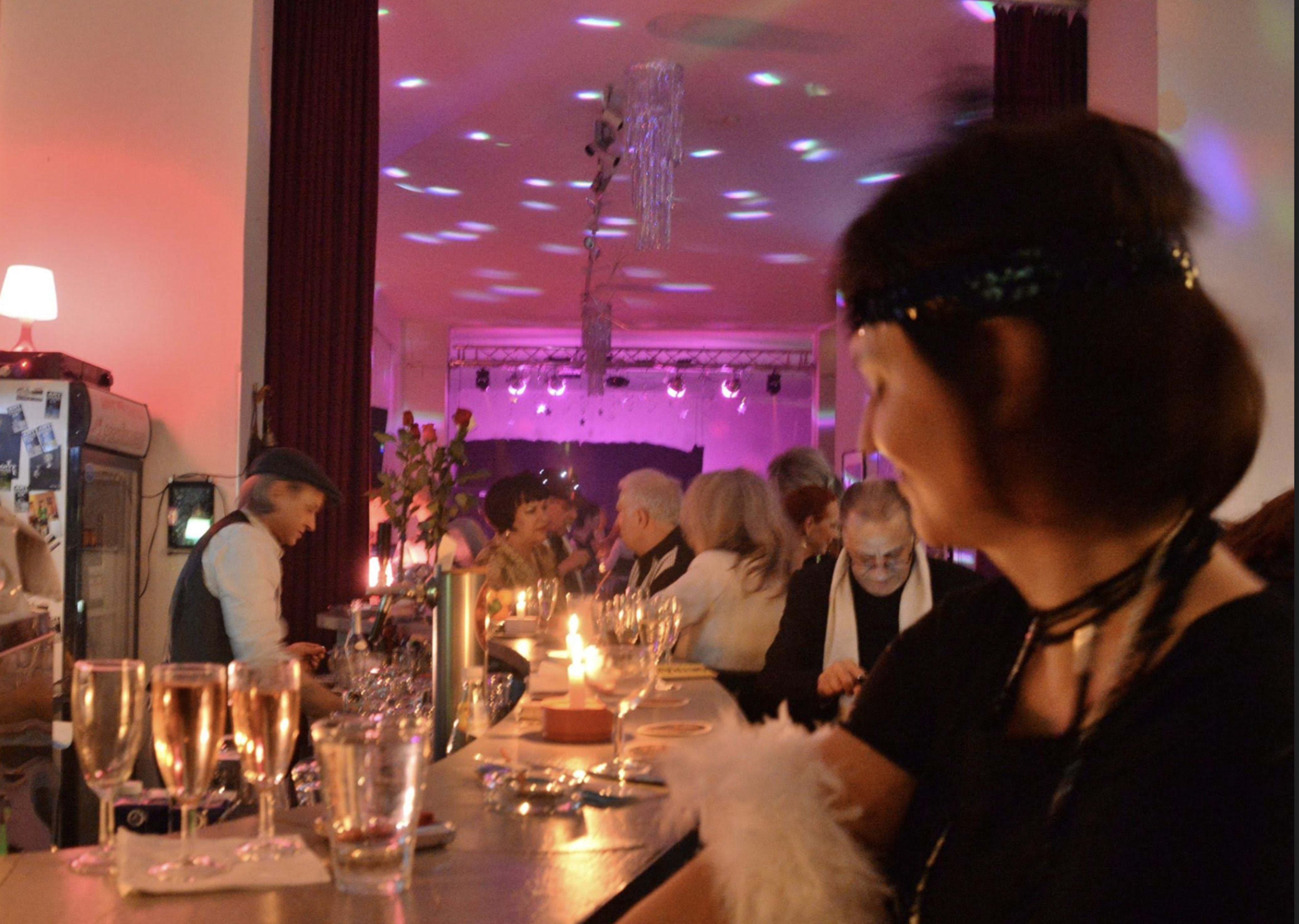 A lively gathering is depicted at a bar, with guests enjoying drinks and colorful lights illuminating the scene.