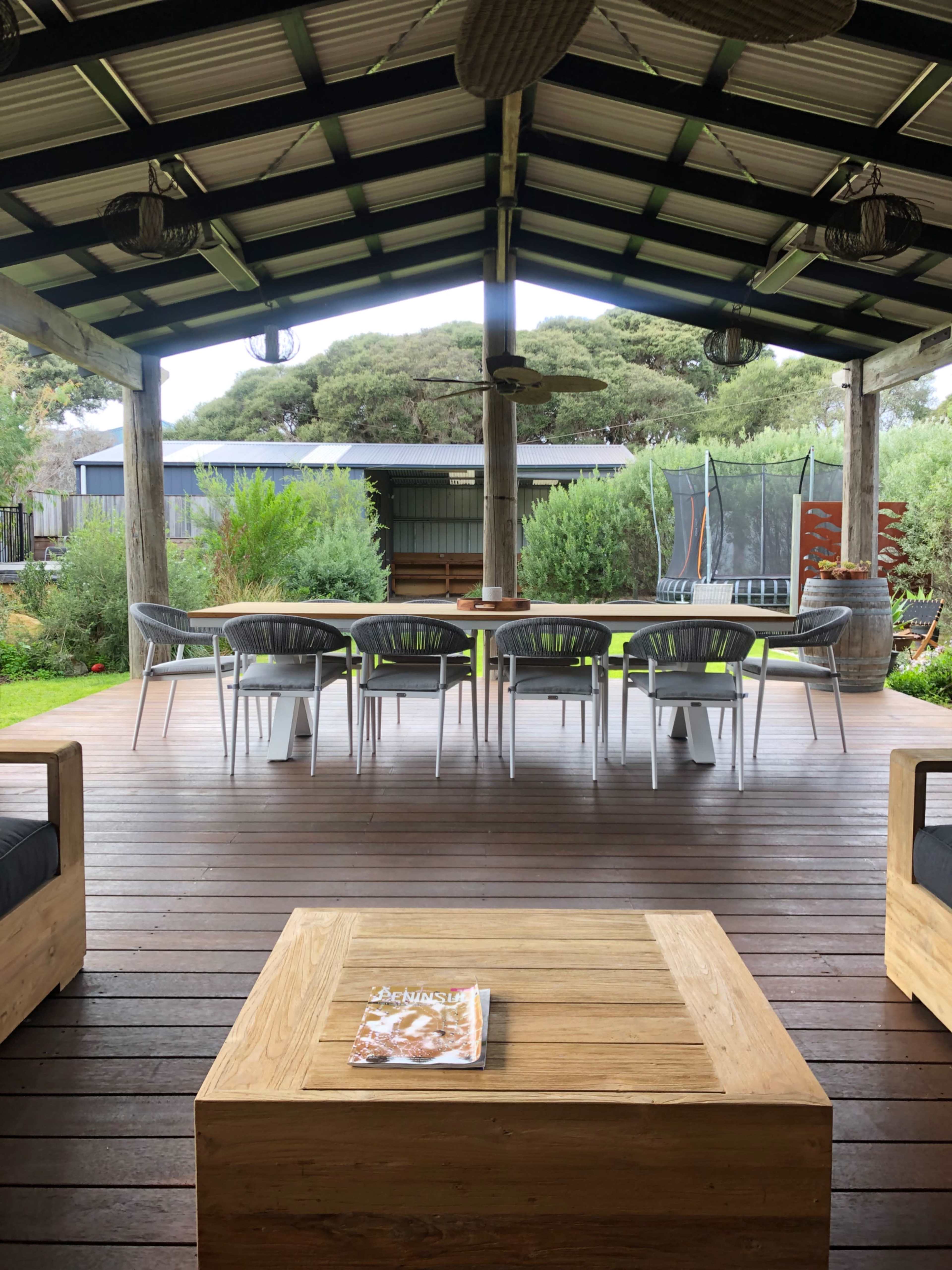 A spacious outdoor seating area features a large table surrounded by chairs under a covered wooden patio, with greenery and a trampoline visible in the background.
