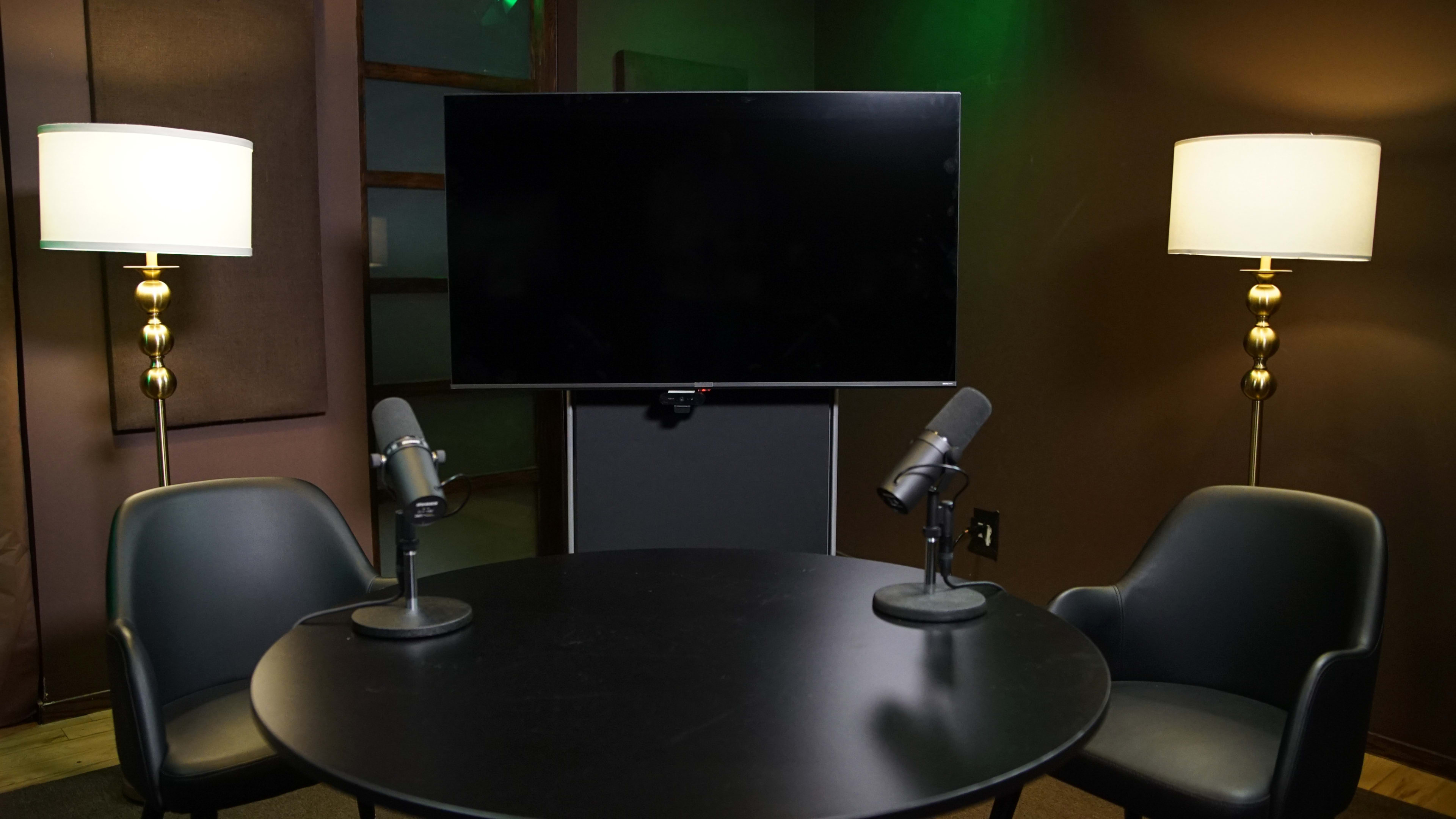 A small studio setup with a black round table, two chairs, a television screen, and two microphones positioned on the table.