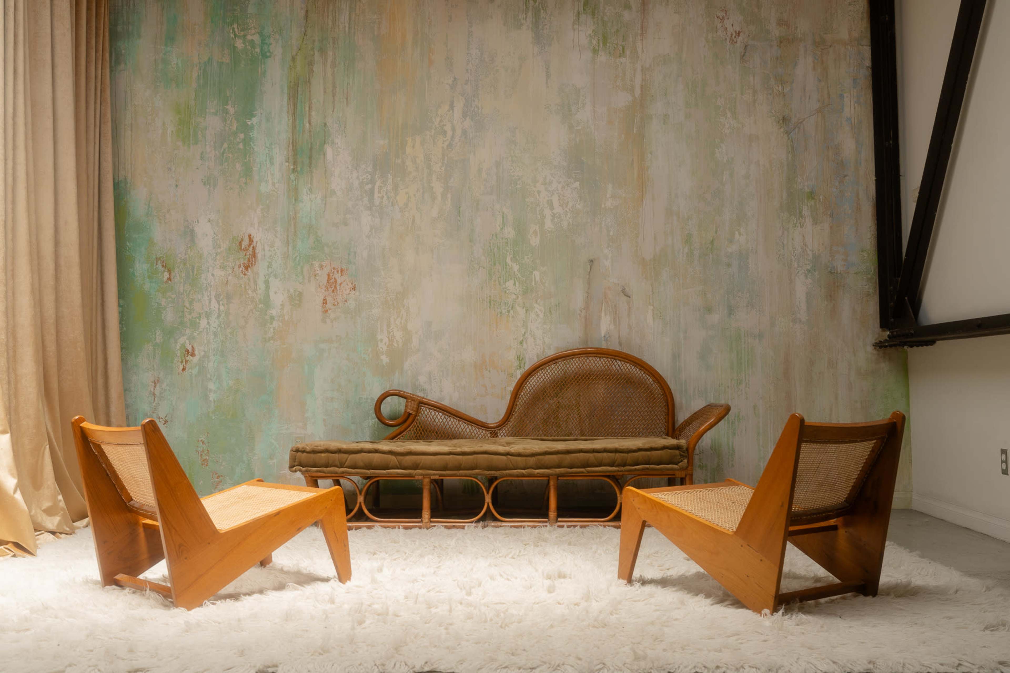 A minimalist seating arrangement with a curved rattan sofa and two angled wooden chairs on a textured rug in a softly colored room.