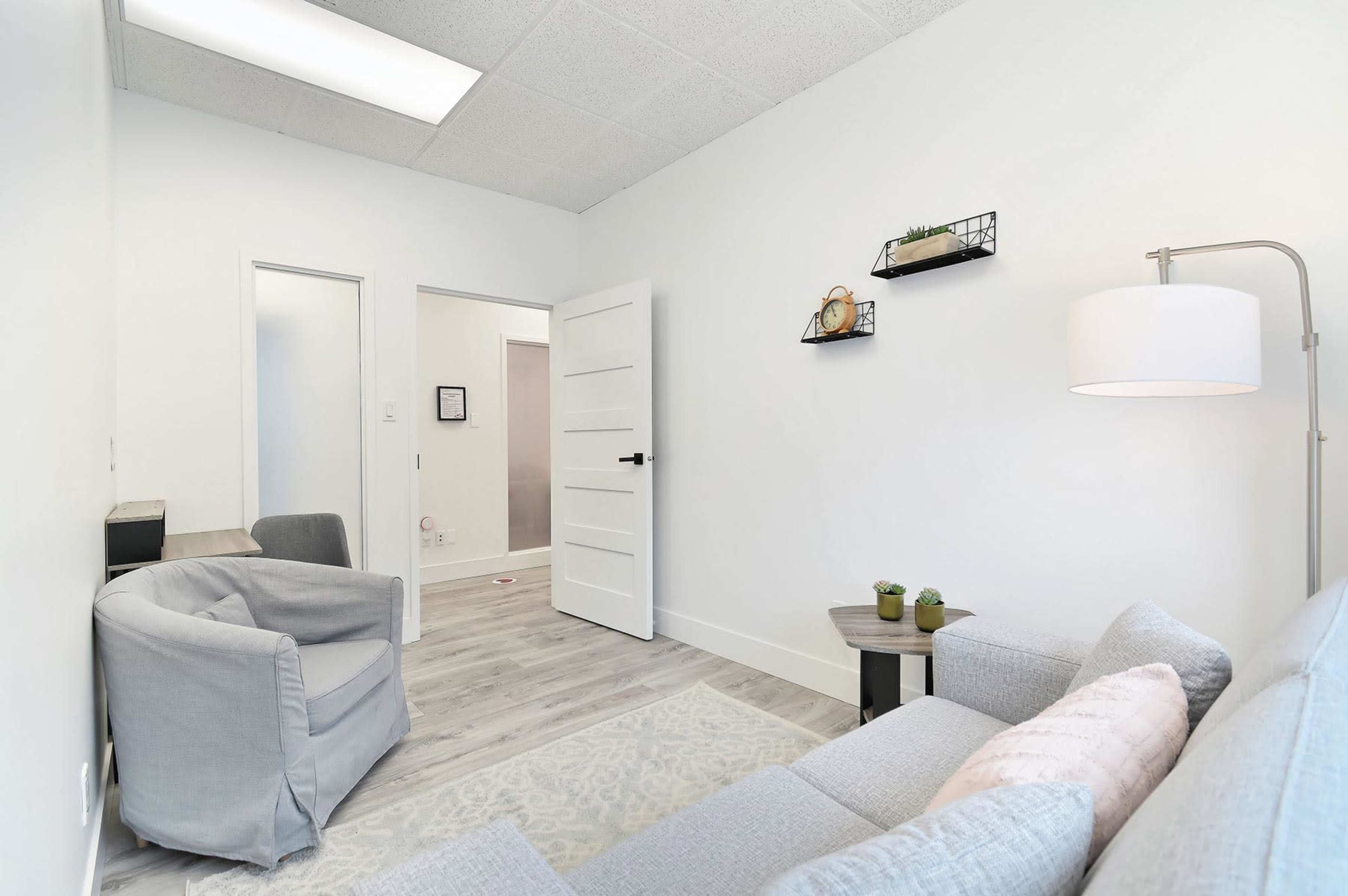 A small, modern waiting room with a gray sofa, a floor lamp, and a table with decorative plants against a white wall.