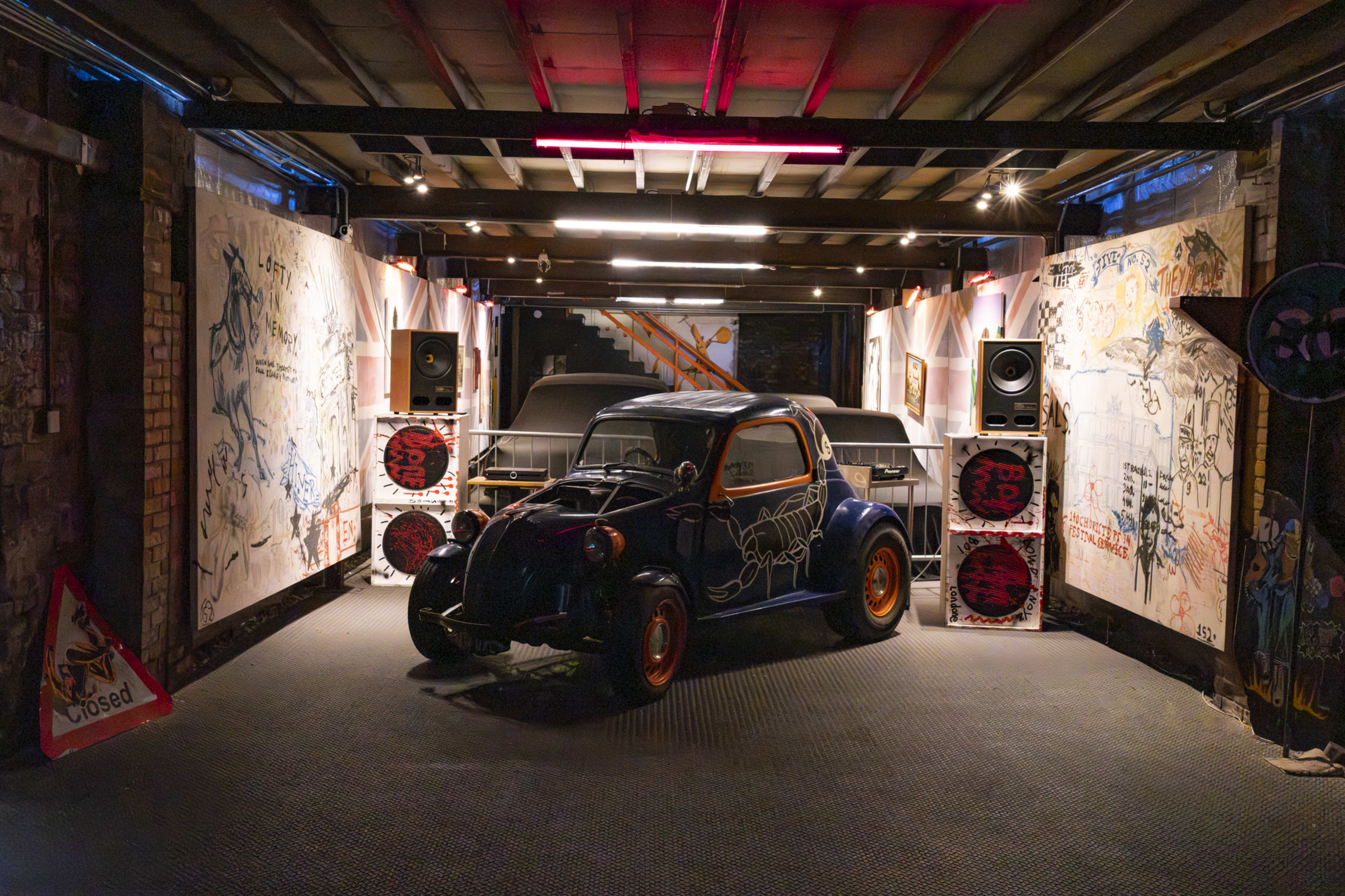 A vintage car is displayed in a graffiti-covered room with speakers and warning signs.