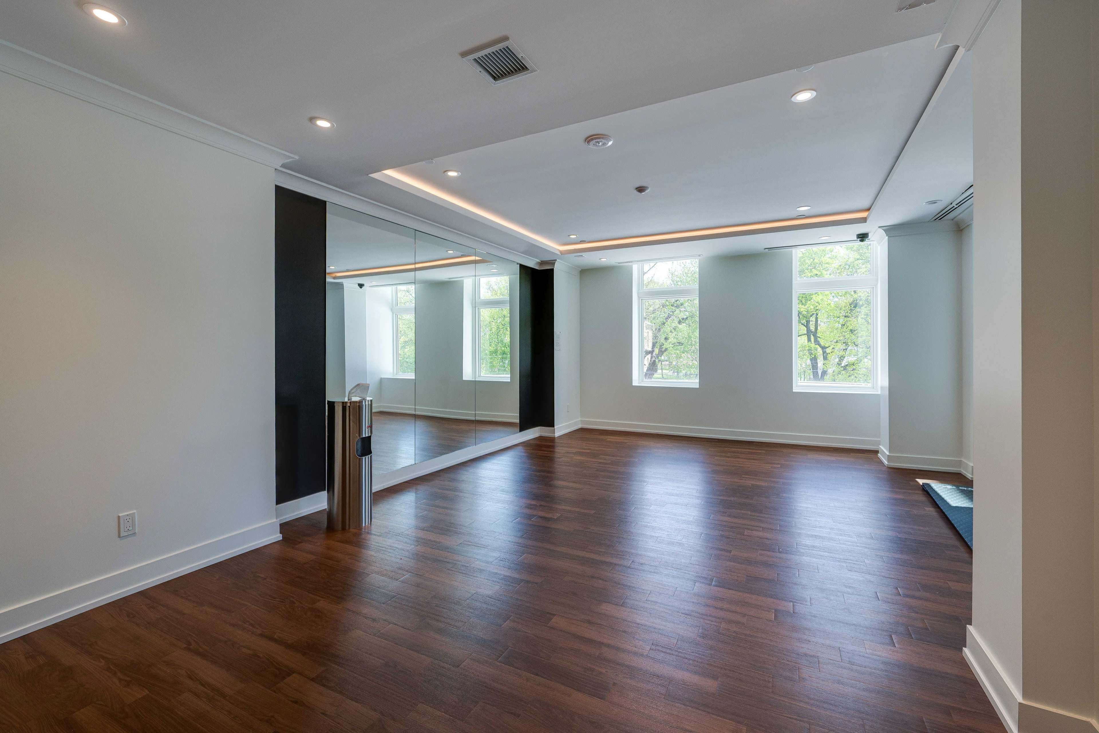 The image shows a spacious, well-lit room with hardwood flooring, large windows, and a mirrored wall.
