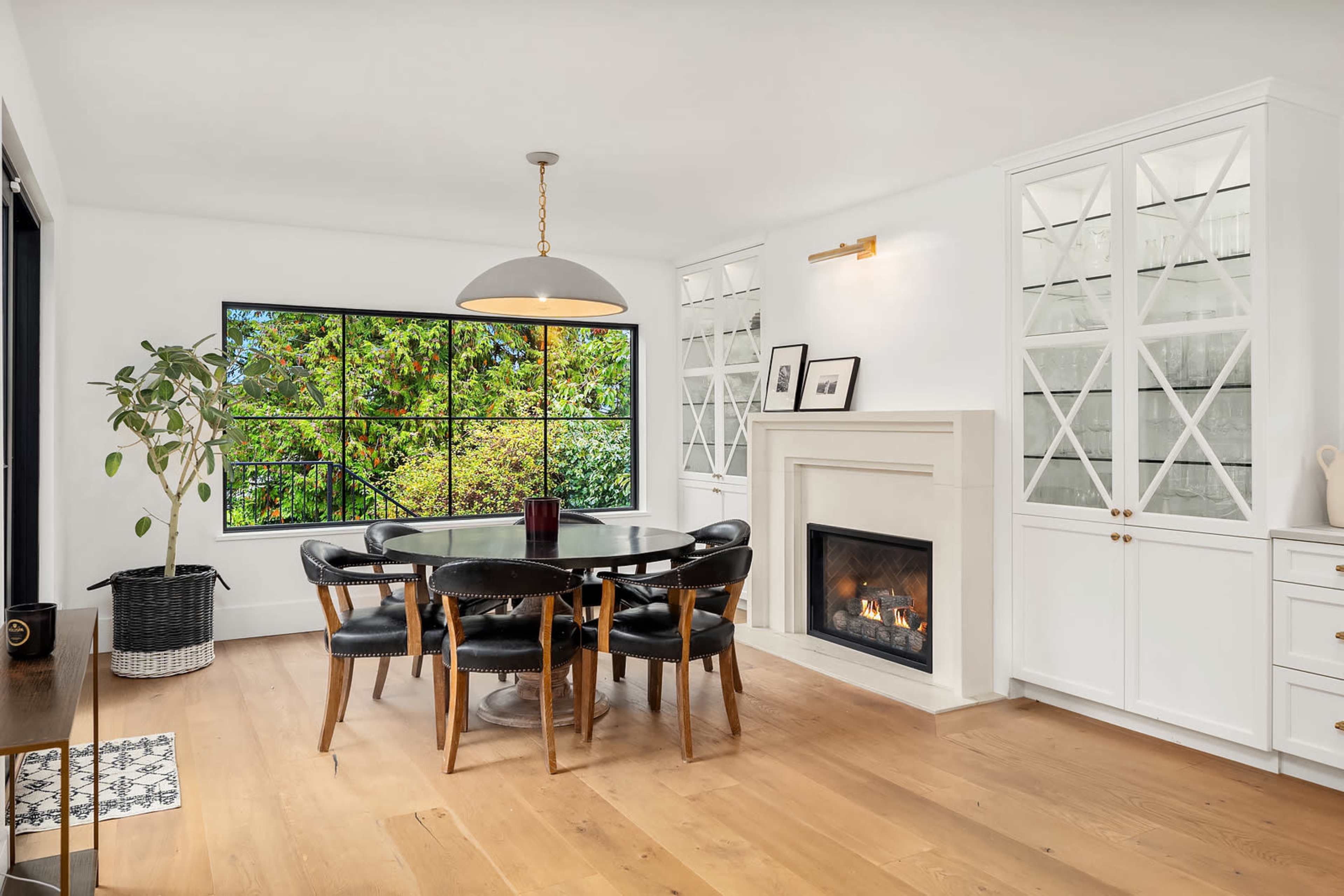 A dining area features a round table surrounded by black chairs, a fireplace, and a large window overlooking greenery.