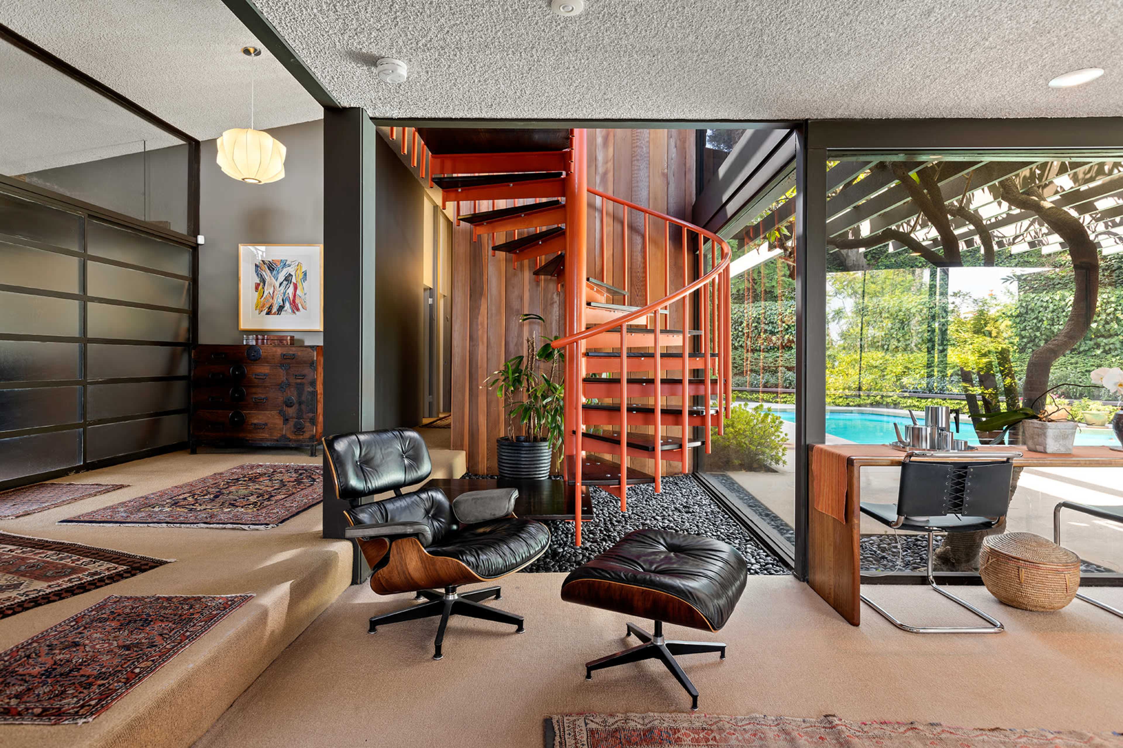 The image shows a modern interior featuring a spiral staircase, a minimalist seating area with an armchair and ottoman, and large windows that offer a view of an outdoor pool and garden.