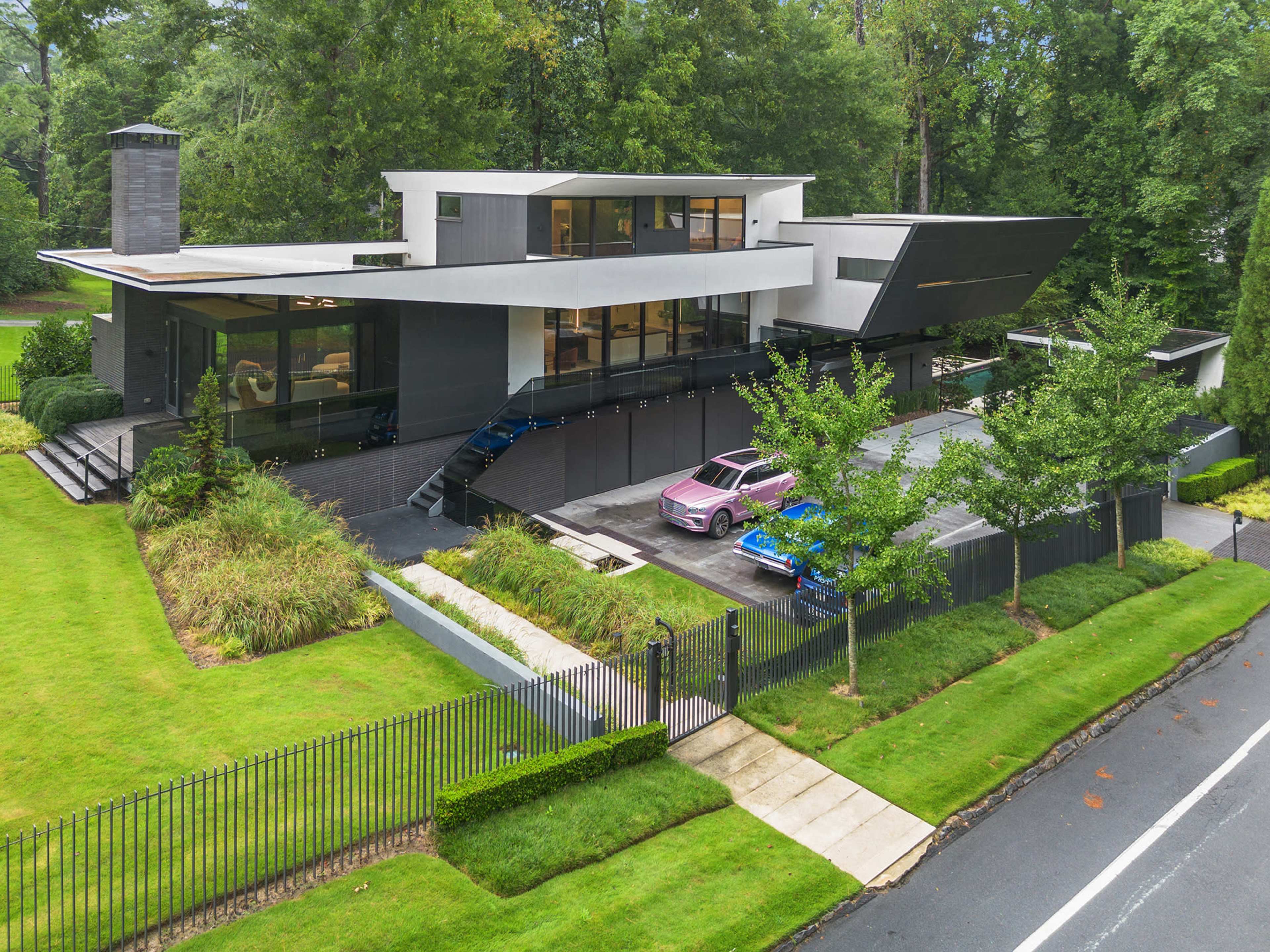 A modern two-story house with a sleek design and large glass windows is surrounded by well-manicured lawns and parked colorful sports cars.